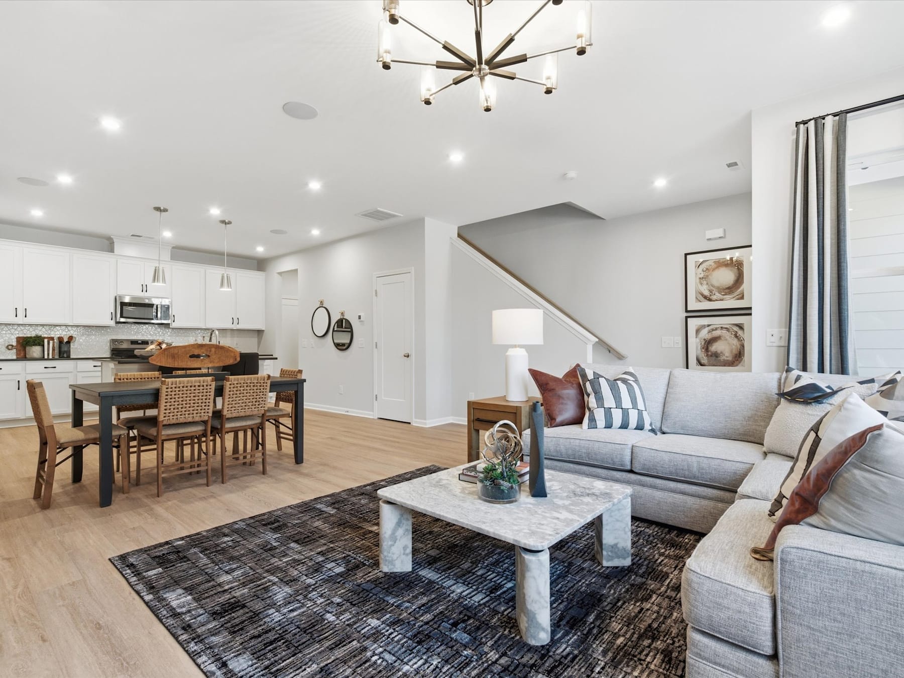 A modern and spacious open-concept living room with a dining area and kitchen, featuring a large gray sofa, a marble coffee table, and a statement light fixture overhead.