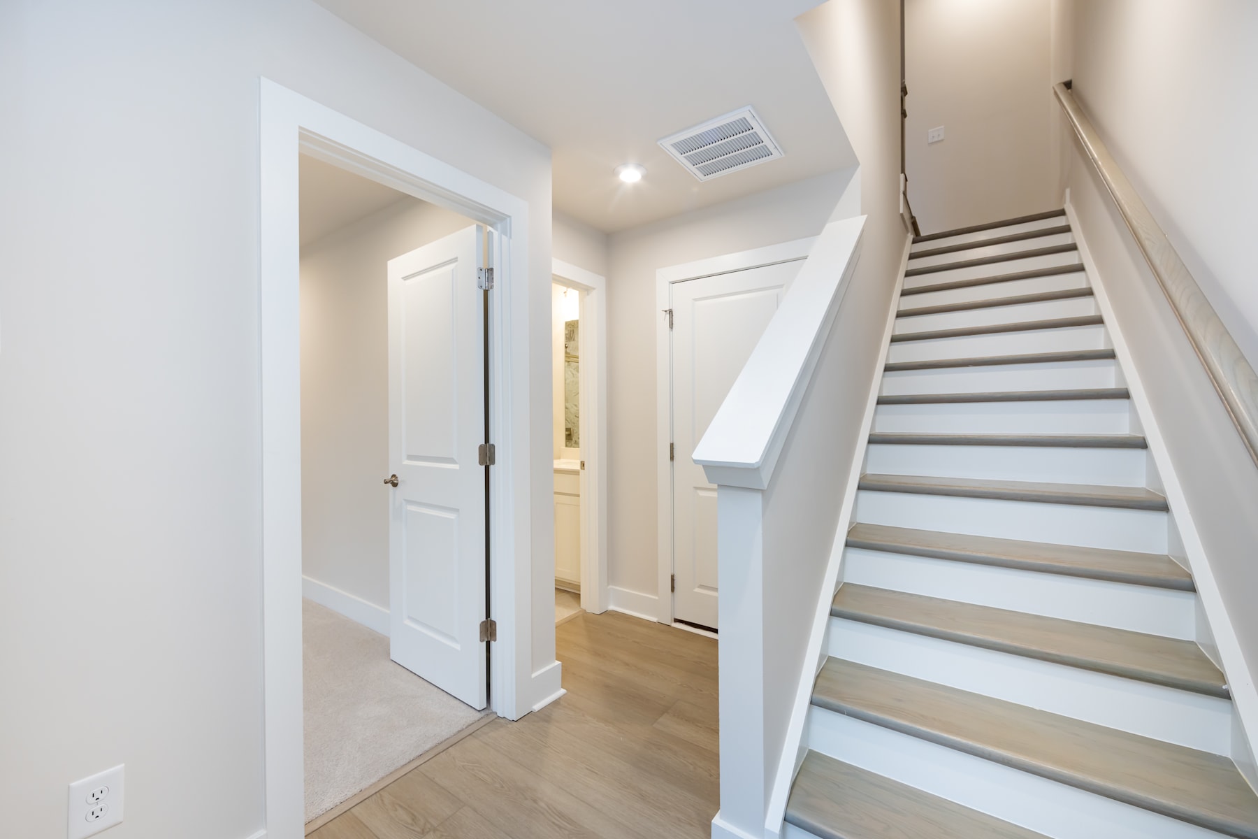 A bright and airy hallway with a staircase leading upstairs, featuring white walls, a wooden floor, and a closed door in the background.