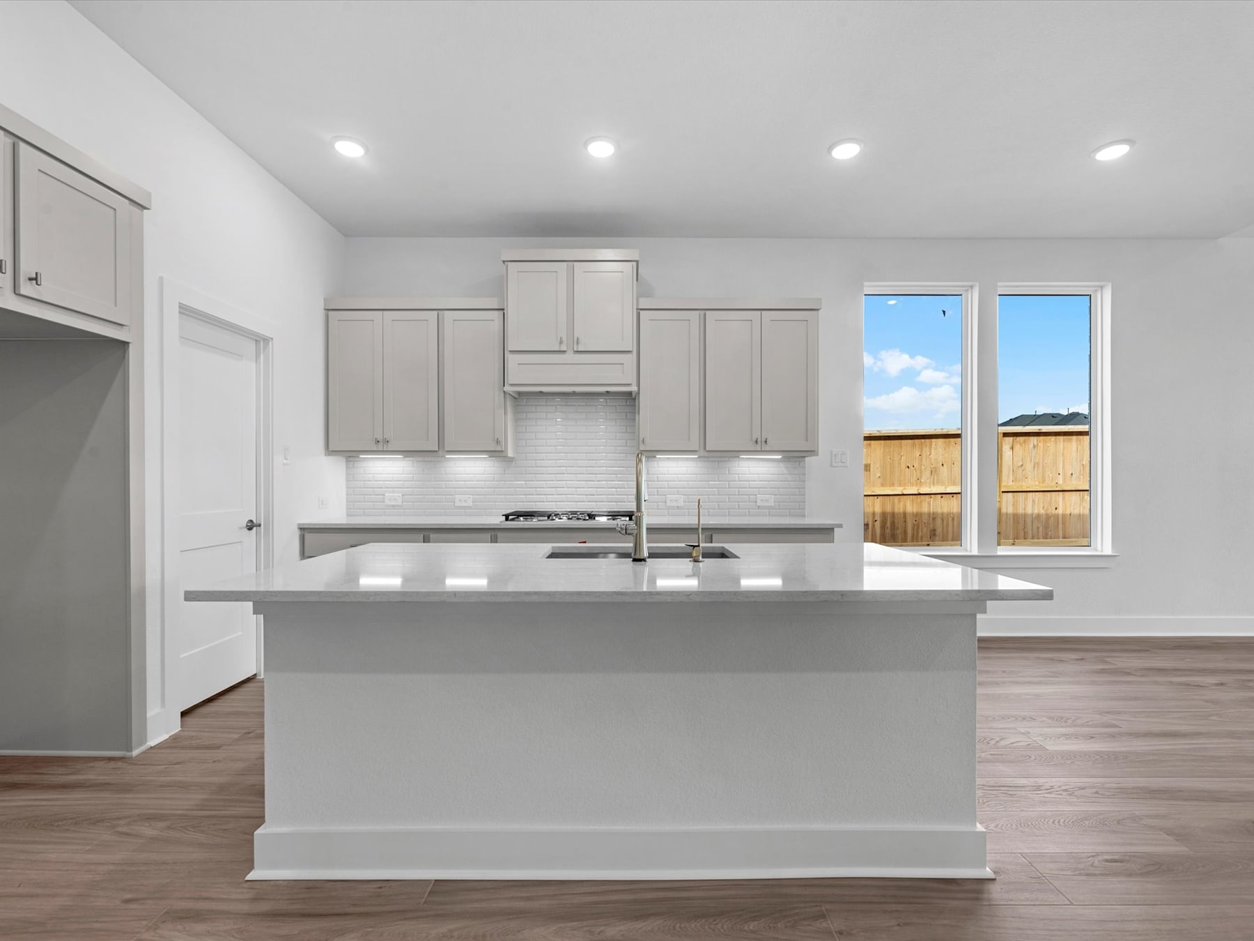A modern, minimalist kitchen with white cabinets, a central island, and large windows overlooking a backyard.