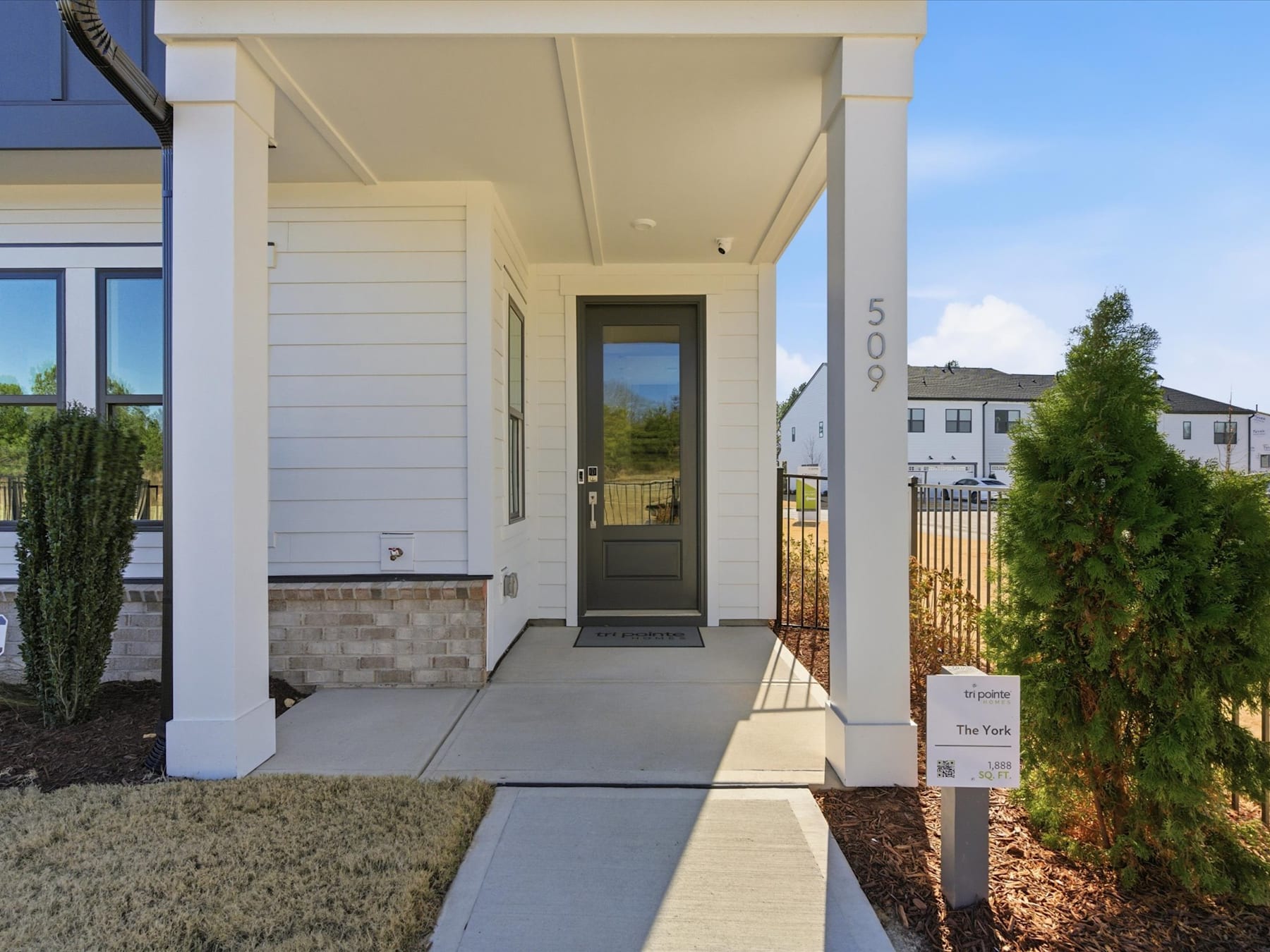 A modern, white-colored house with a covered porch and a walkway leading to the front door, surrounded by landscaping and other residential buildings in the background.