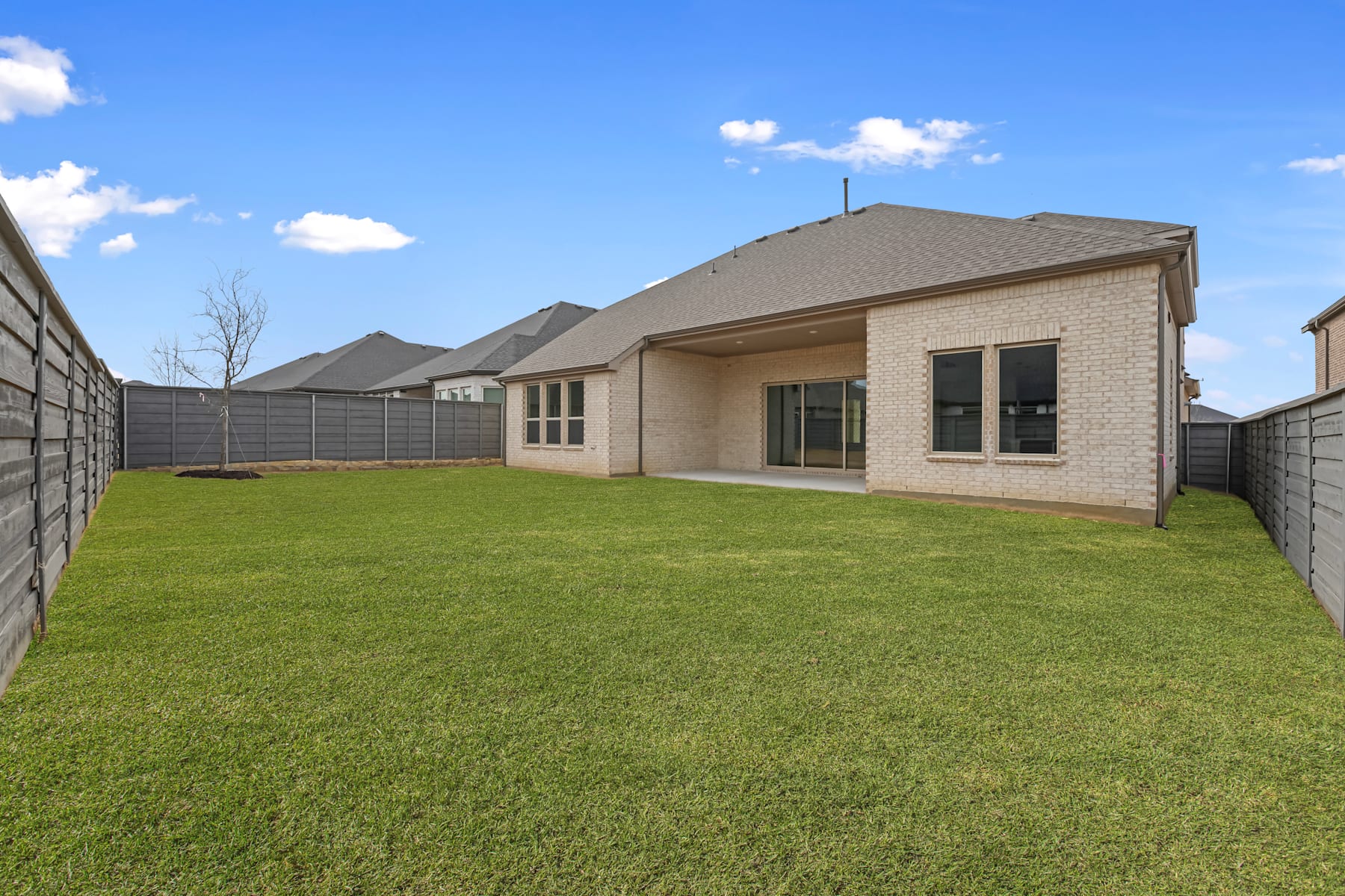 A well-manicured lawn surrounds a modern, single-story brick house with a tiled roof, set against a clear blue sky with scattered clouds.