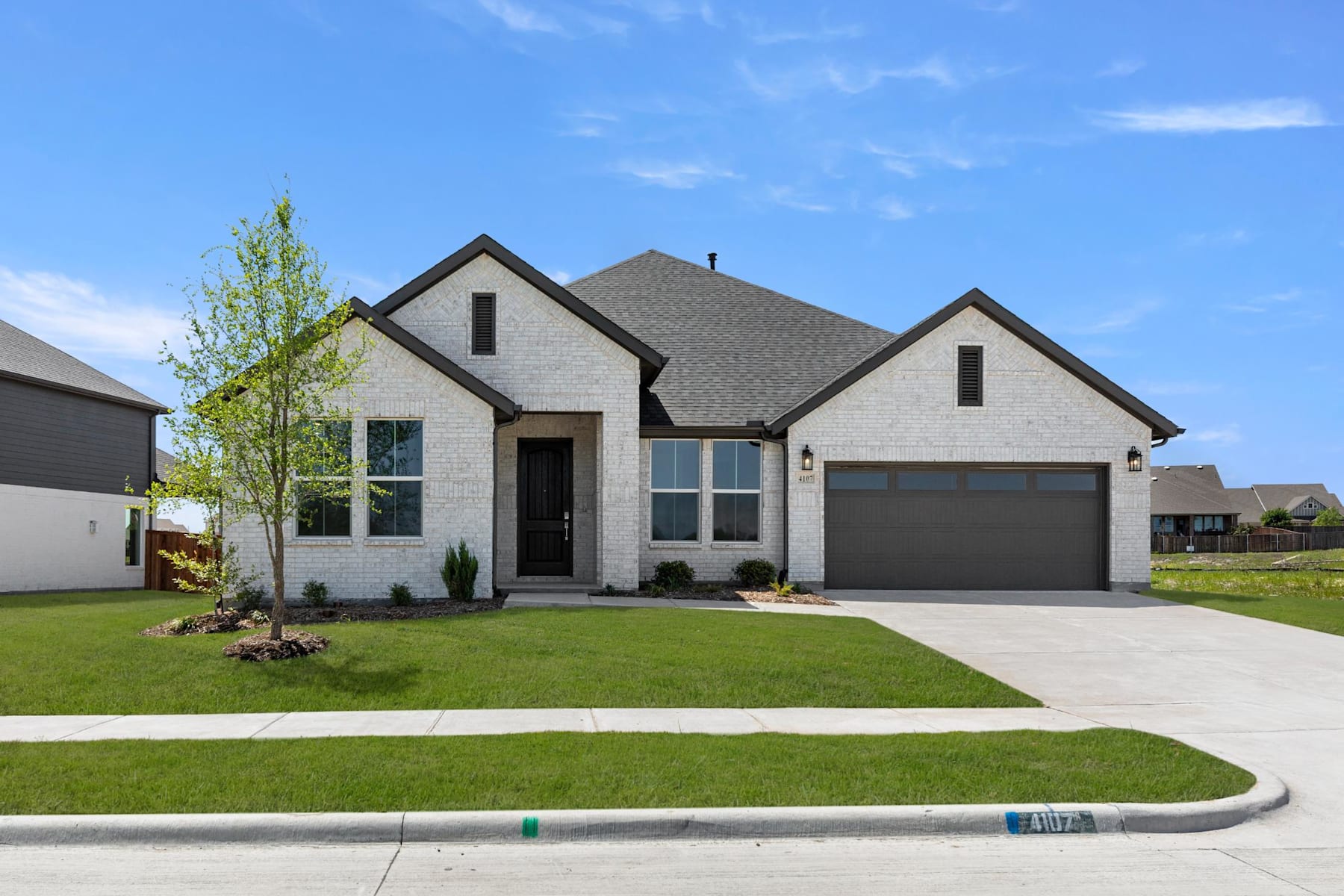 A modern two-story house with a gray roof and white exterior stands on a well-manicured lawn, surrounded by a clear blue sky with scattered clouds.