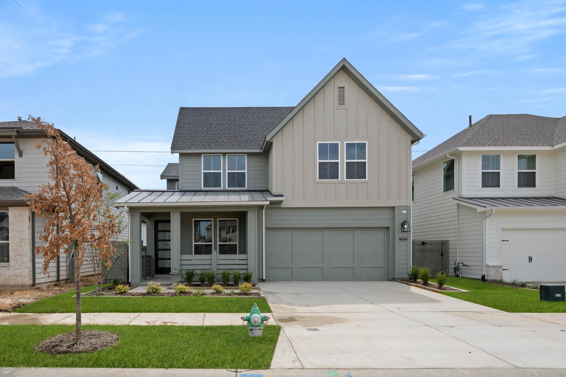 A two-story residential house with a gray roof, beige siding, and a garage door stands in the foreground, surrounded by a grassy lawn and other houses in the background.