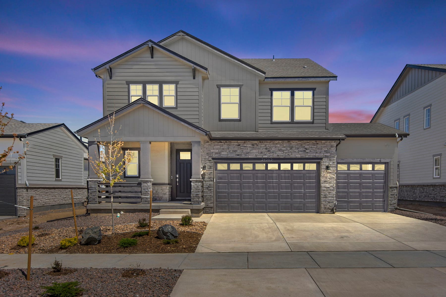A two-story residential house with a stone facade, a garage, and a paved driveway in the foreground, set against a backdrop of a clear blue sky with a colorful sunset.