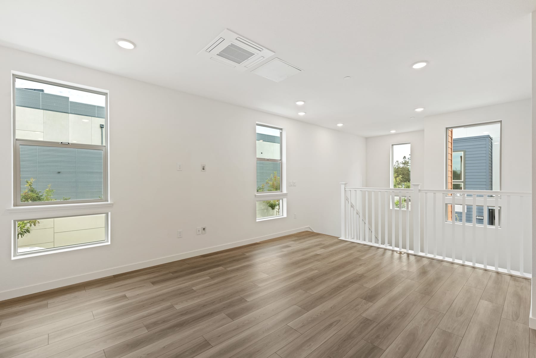 An empty, well-lit room with hardwood floors, white walls, and large windows providing natural light. The room appears to be a bedroom or living space with a staircase visible in the background.