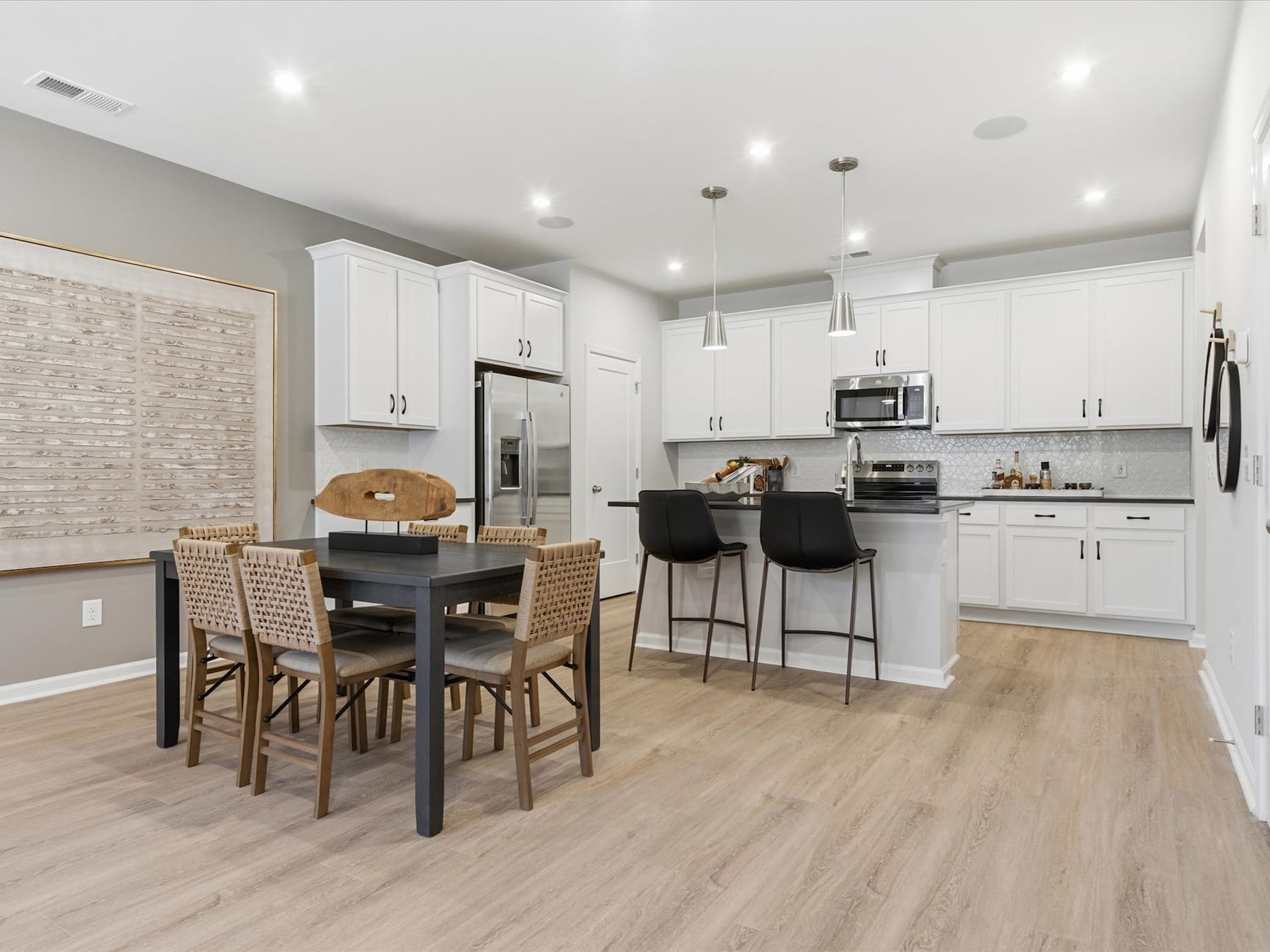 A modern, open-concept kitchen and dining area with white cabinets, stainless steel appliances, a wooden dining table, and wicker bar stools.