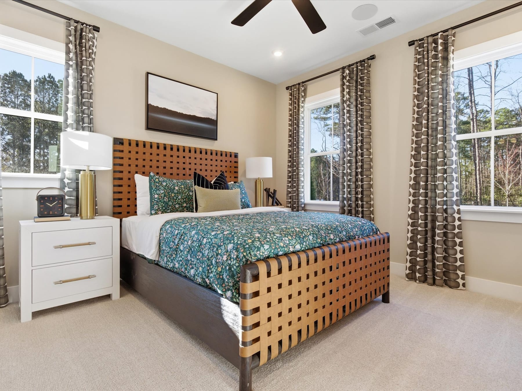 A cozy and well-furnished bedroom with a patterned bedspread, wooden headboard, and large windows overlooking a wooded outdoor scene.