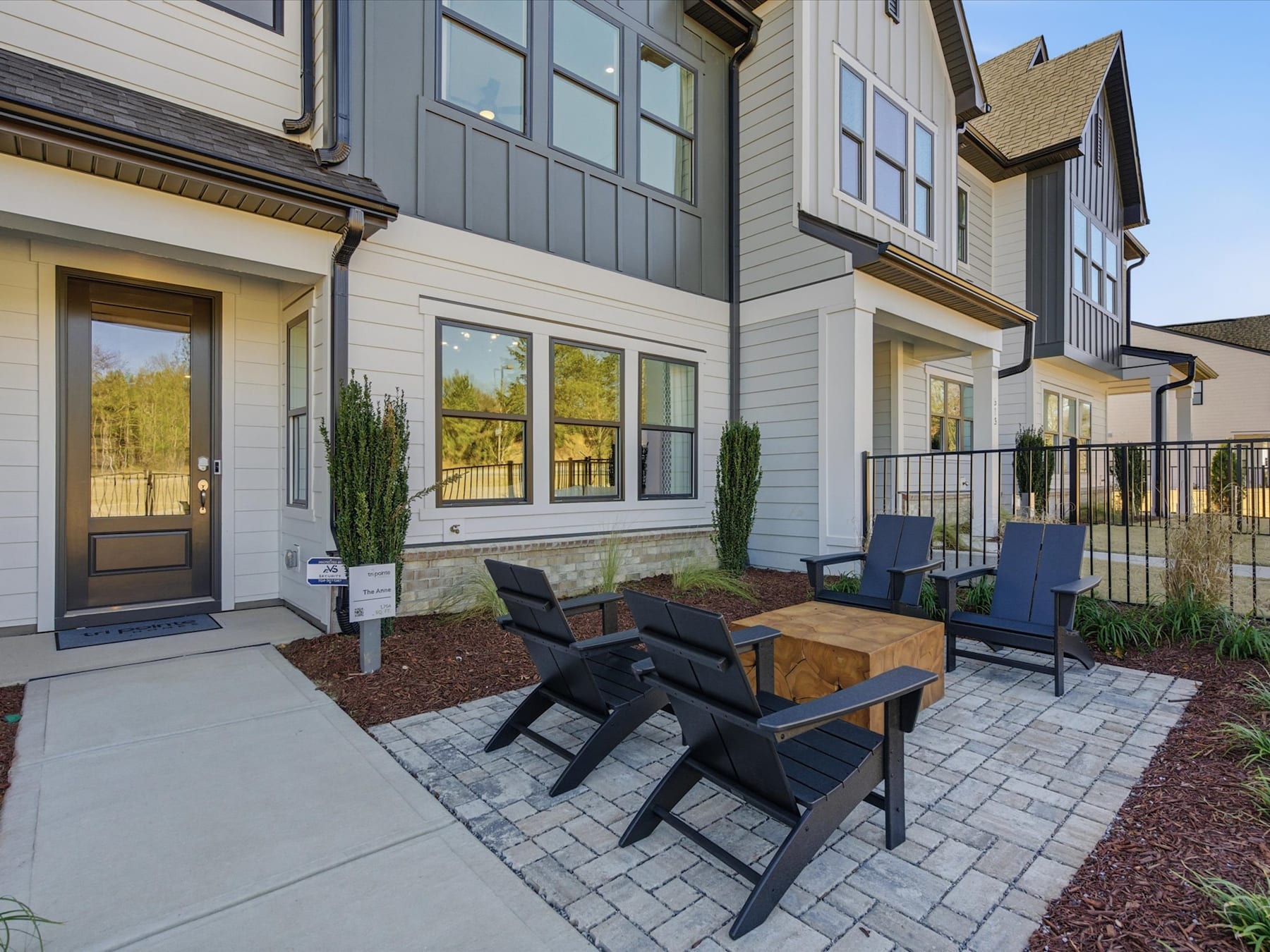 A cozy outdoor seating area with black wooden chairs and a table, surrounded by a paved patio and landscaping, in front of a multi-story residential building with gray siding and large windows.