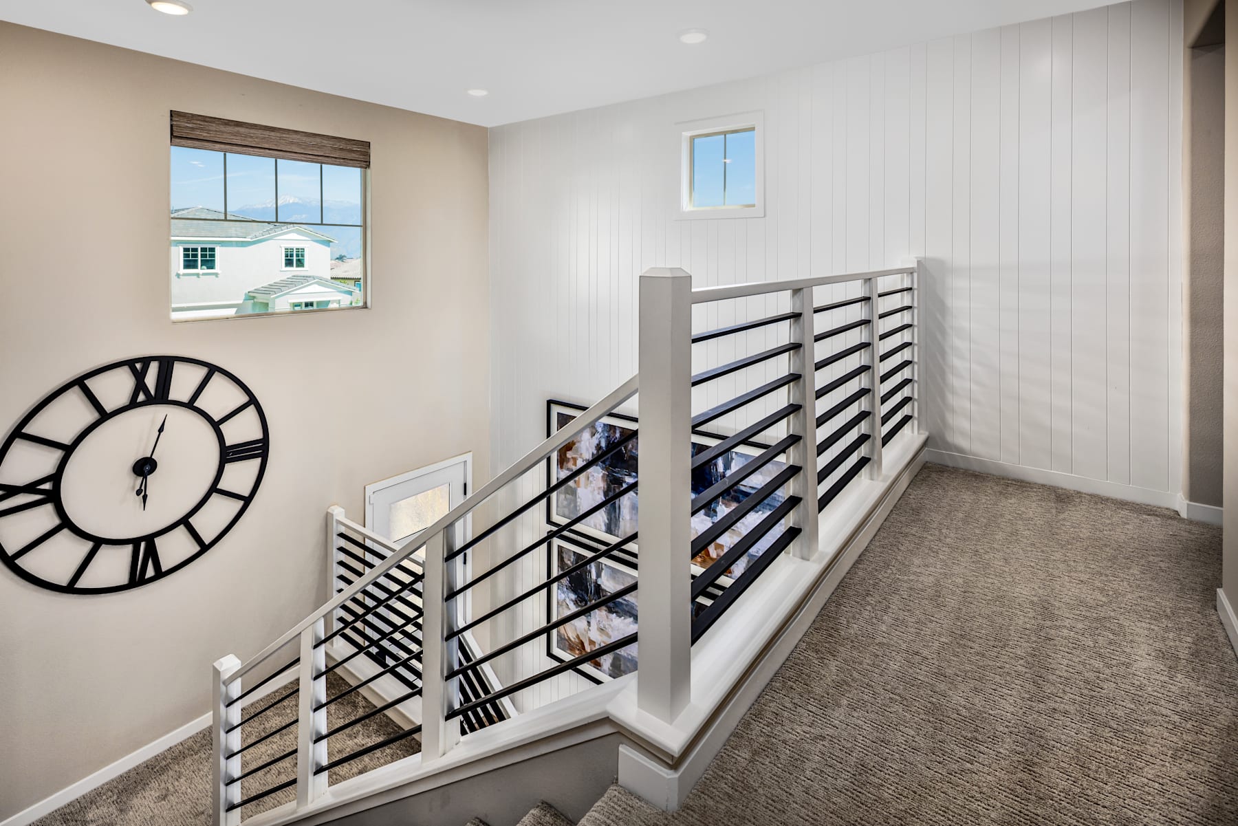 A modern staircase with a large clock on the wall, leading up to a second floor with a window overlooking a residential neighborhood.