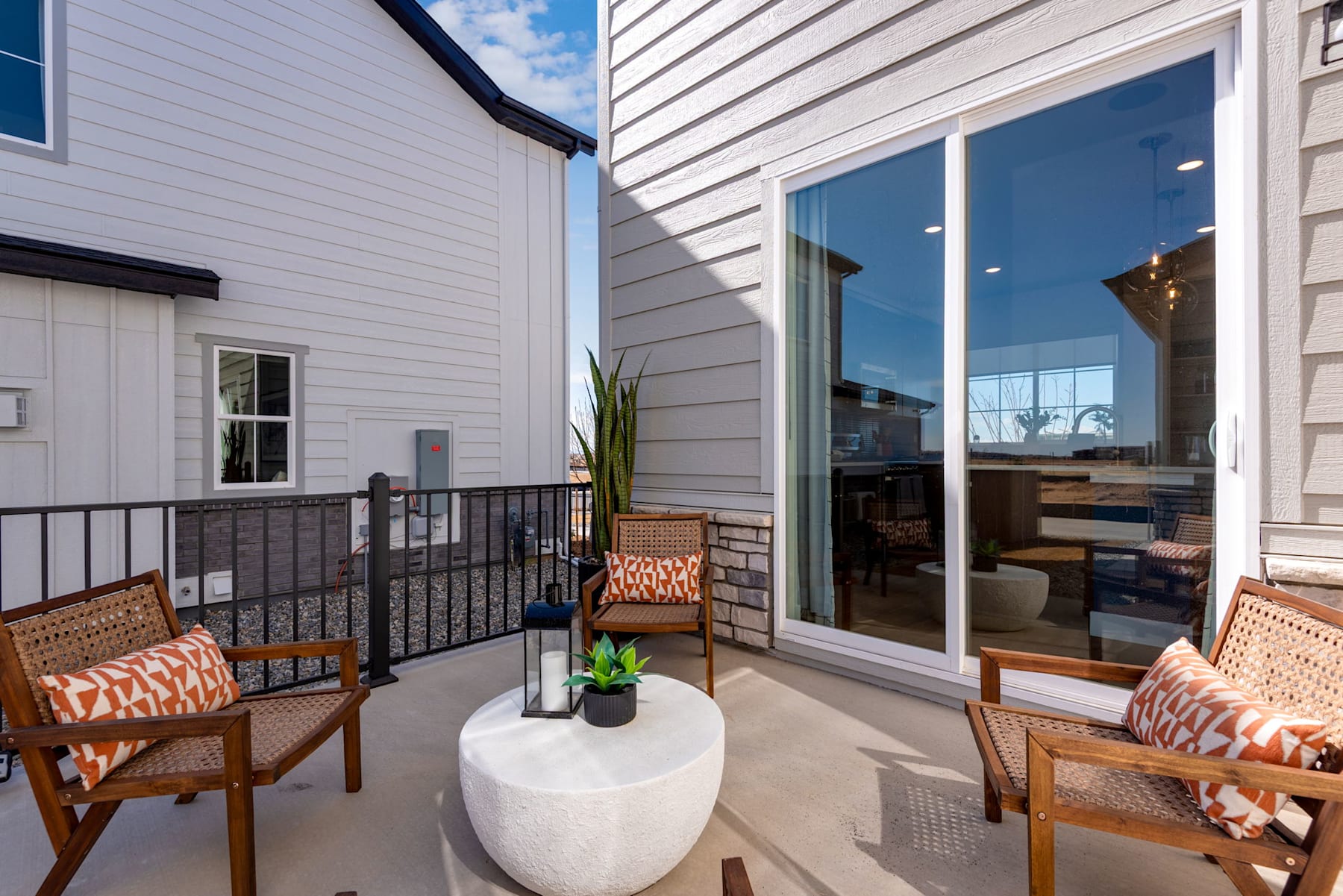 A cozy outdoor patio with wicker furniture, a potted plant, and a glass sliding door leading to an interior space with visible shelving and decor.
