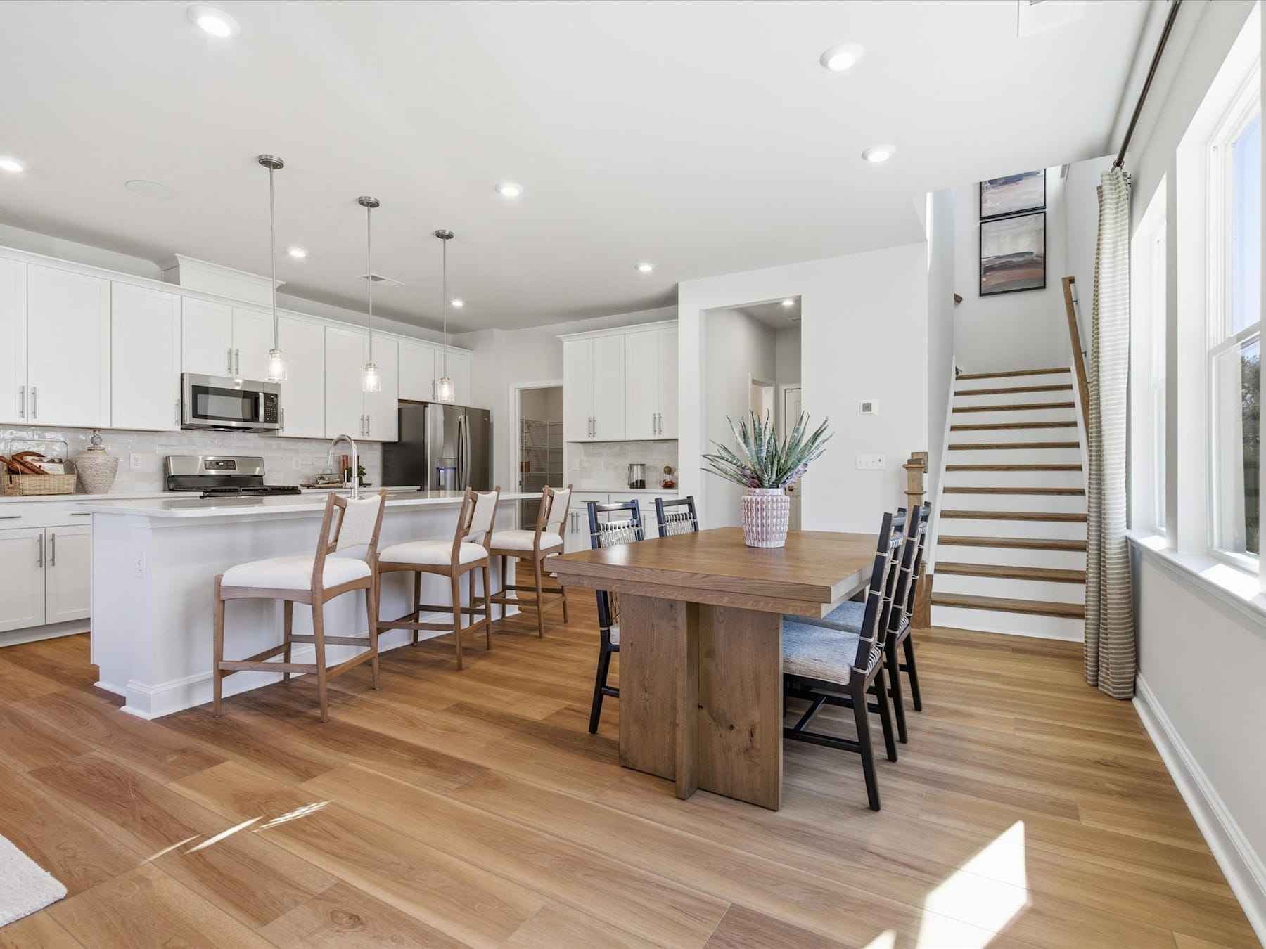A modern, open-concept kitchen and dining area with white cabinets, wooden floors, and a large wooden dining table surrounded by chairs.
