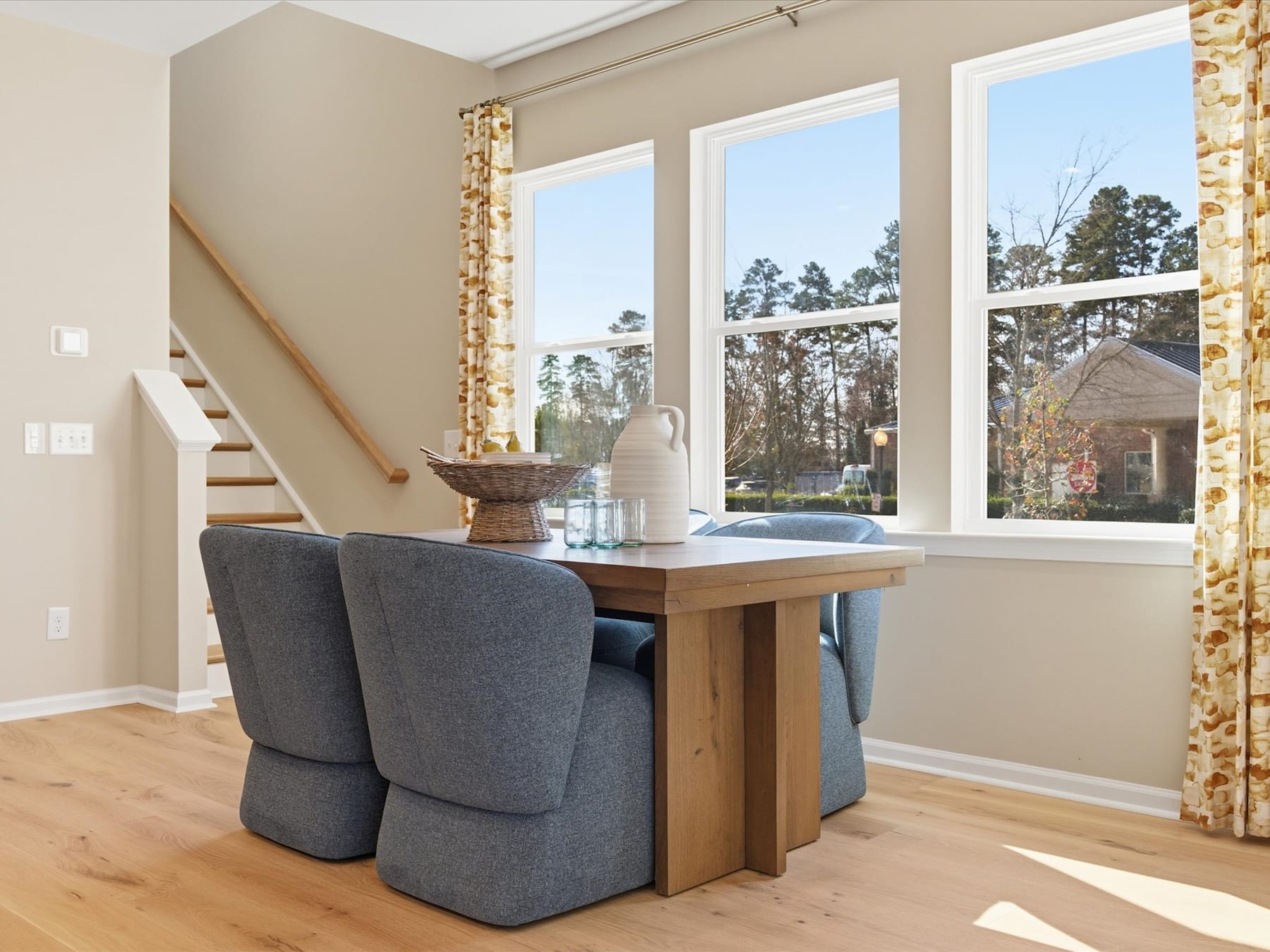 A cozy dining area with a wooden table and gray upholstered chairs, surrounded by large windows overlooking a wooded landscape outside.