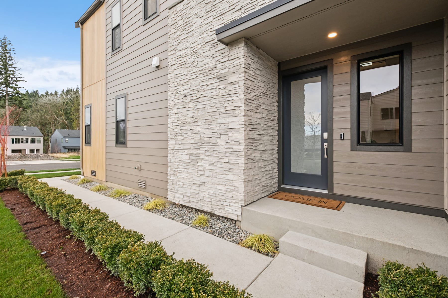A modern, two-story residential building with a stone facade, a covered entryway, and a well-landscaped front yard featuring shrubs and a paved walkway.