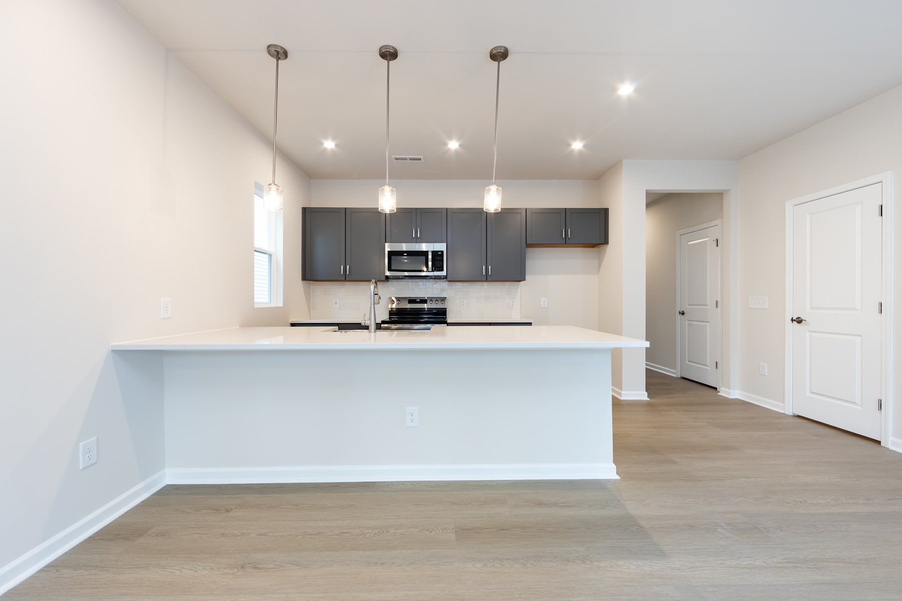 A modern, open-concept kitchen with gray cabinets, a white countertop, and pendant lighting fixtures, set against a light-colored hardwood floor.