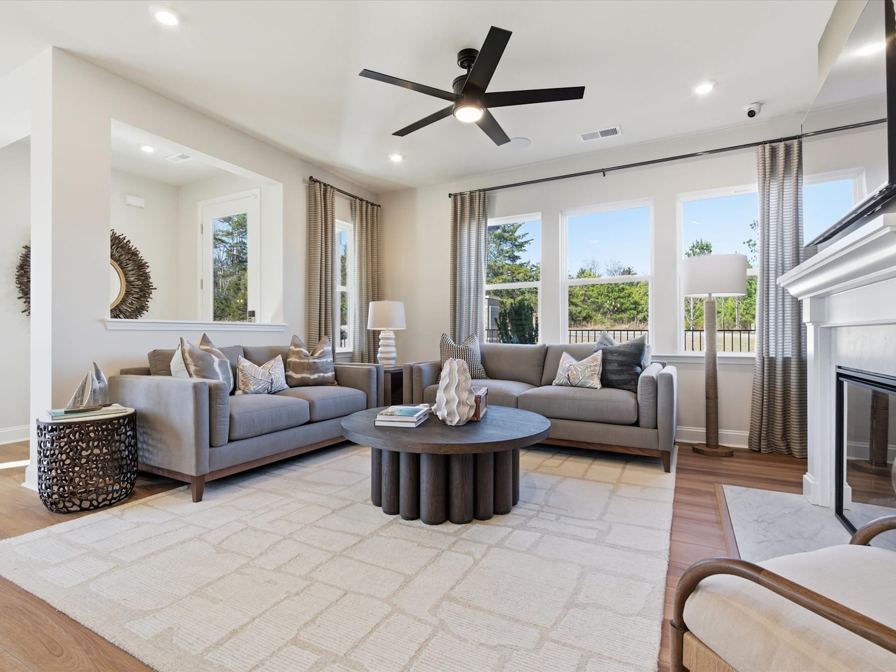 A spacious and well-furnished living room with comfortable gray sofas, a round coffee table, and a ceiling fan, surrounded by large windows offering a scenic outdoor view.