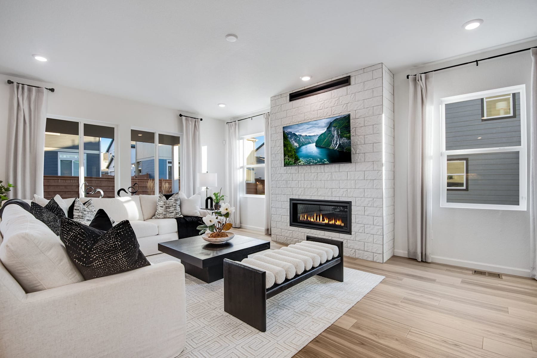 A modern and cozy living room with a fireplace, large windows, and a stylish furniture arrangement featuring a white sofa, black coffee table, and a vibrant landscape artwork on the wall.