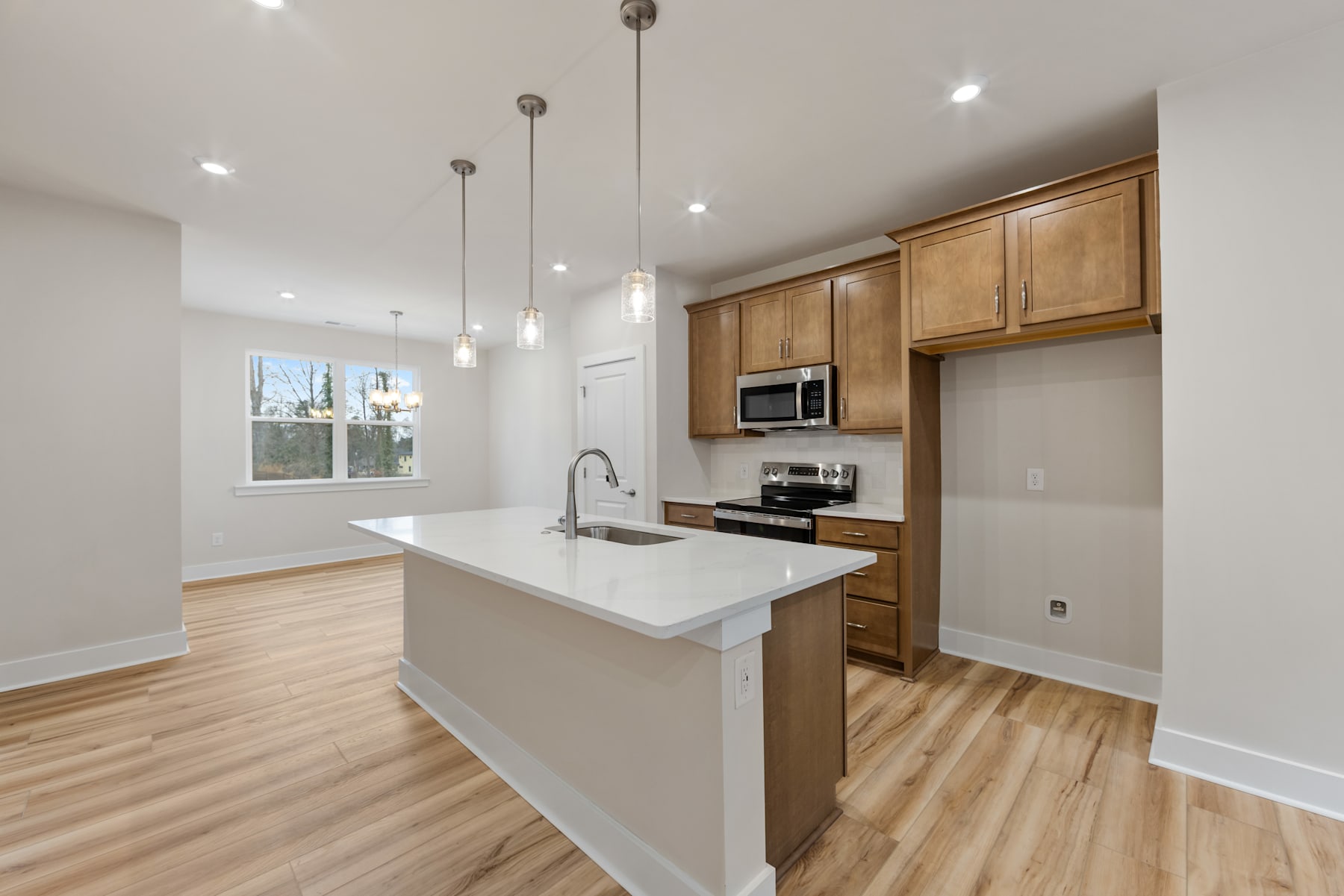 A modern, open-concept kitchen with light-colored wood cabinets, a white countertop, and pendant lighting fixtures hanging from the ceiling.