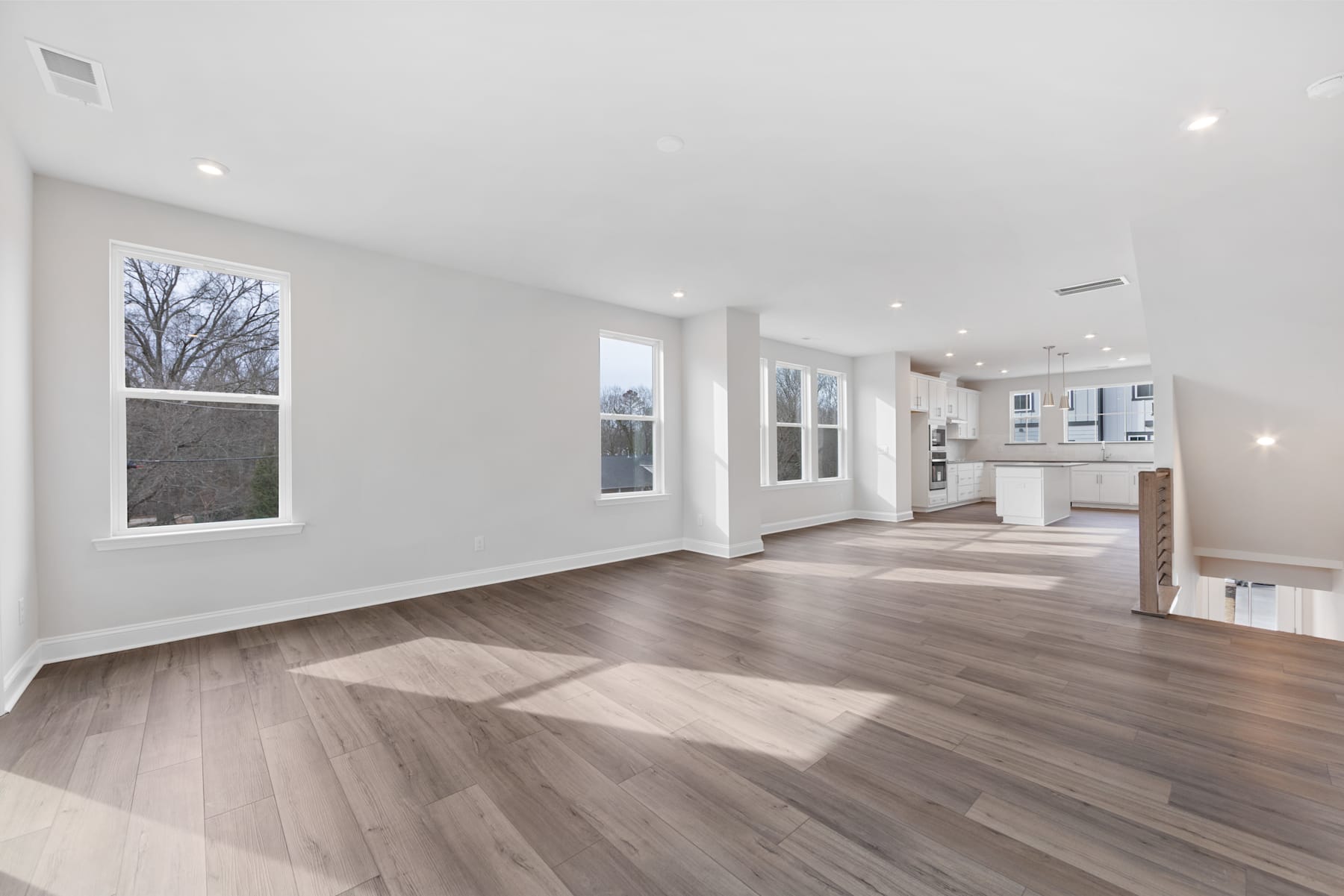 A spacious, open-concept living area with hardwood floors, large windows, and a kitchen visible in the background.