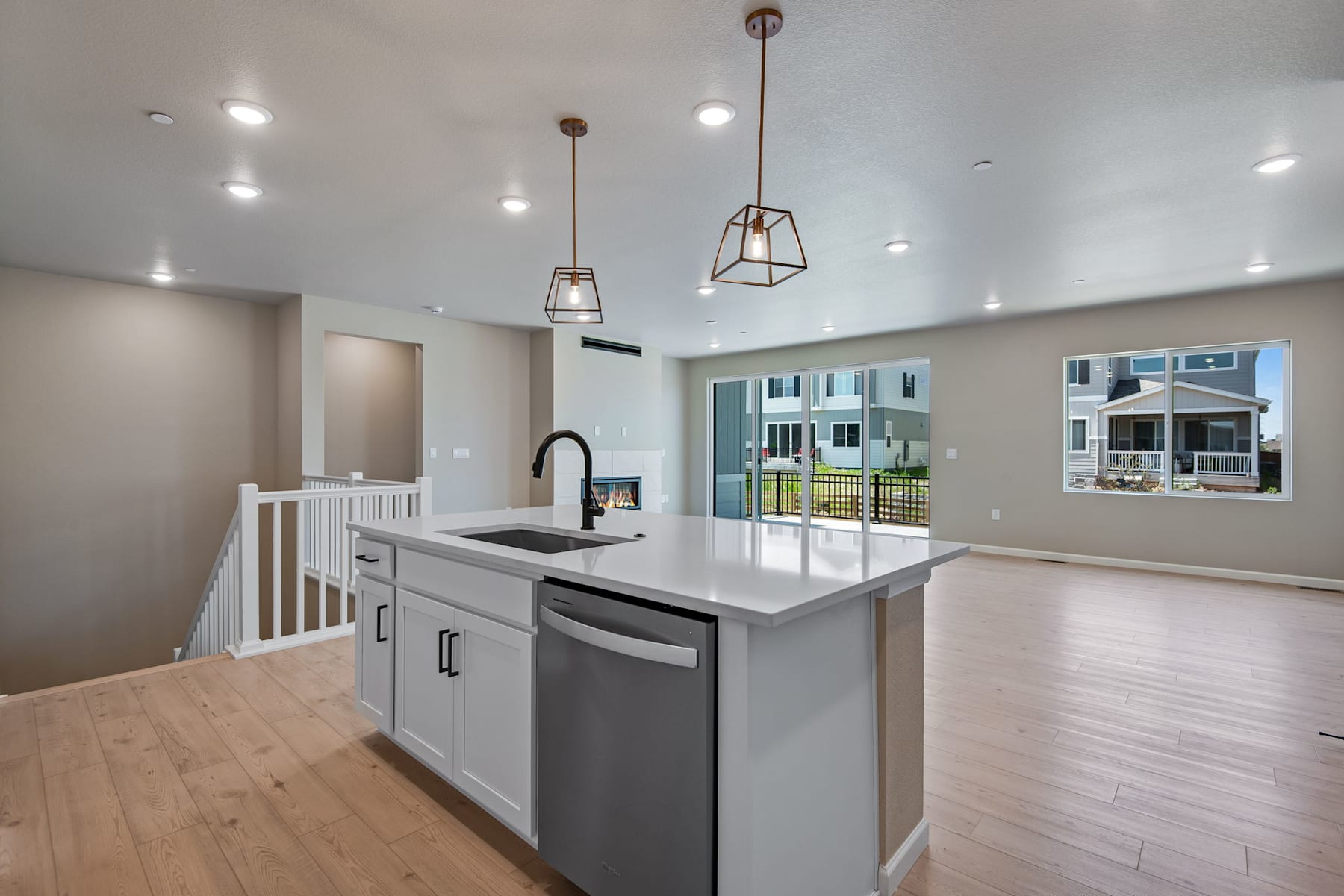 A modern and spacious kitchen with a central island, sleek gray cabinets, and pendant lighting fixtures, set against a backdrop of a residential neighborhood visible through a window.