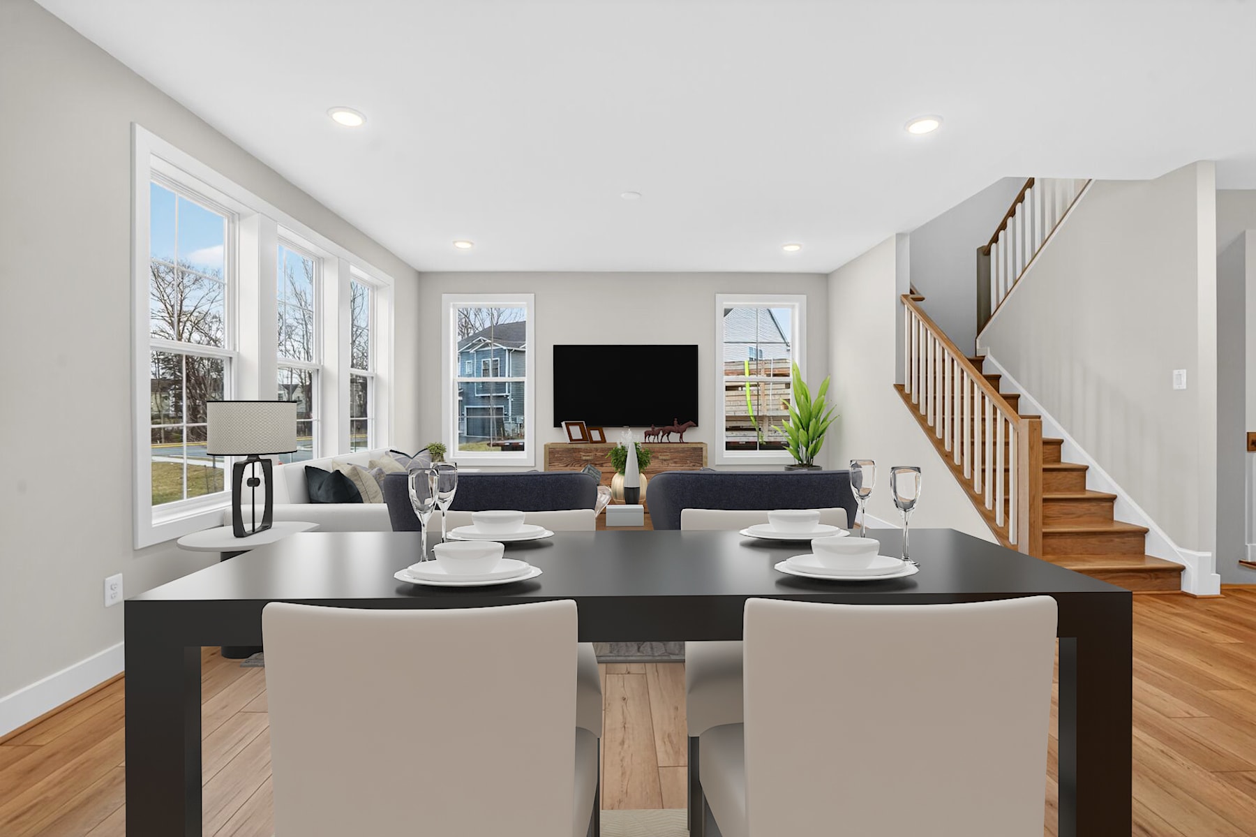 A modern and spacious open-concept living room with a large dining table, comfortable seating, and a television mounted on the wall, surrounded by large windows that let in natural light.