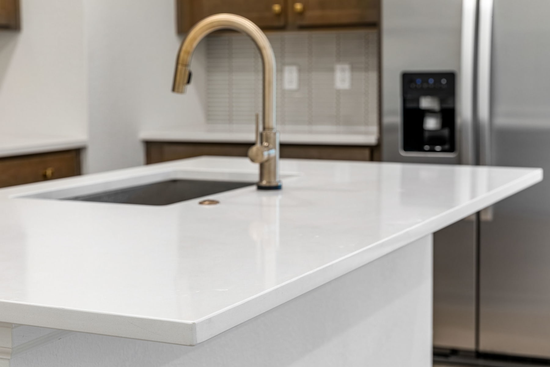 A modern, minimalist kitchen with a white countertop, a stainless steel faucet, and a refrigerator visible in the background.