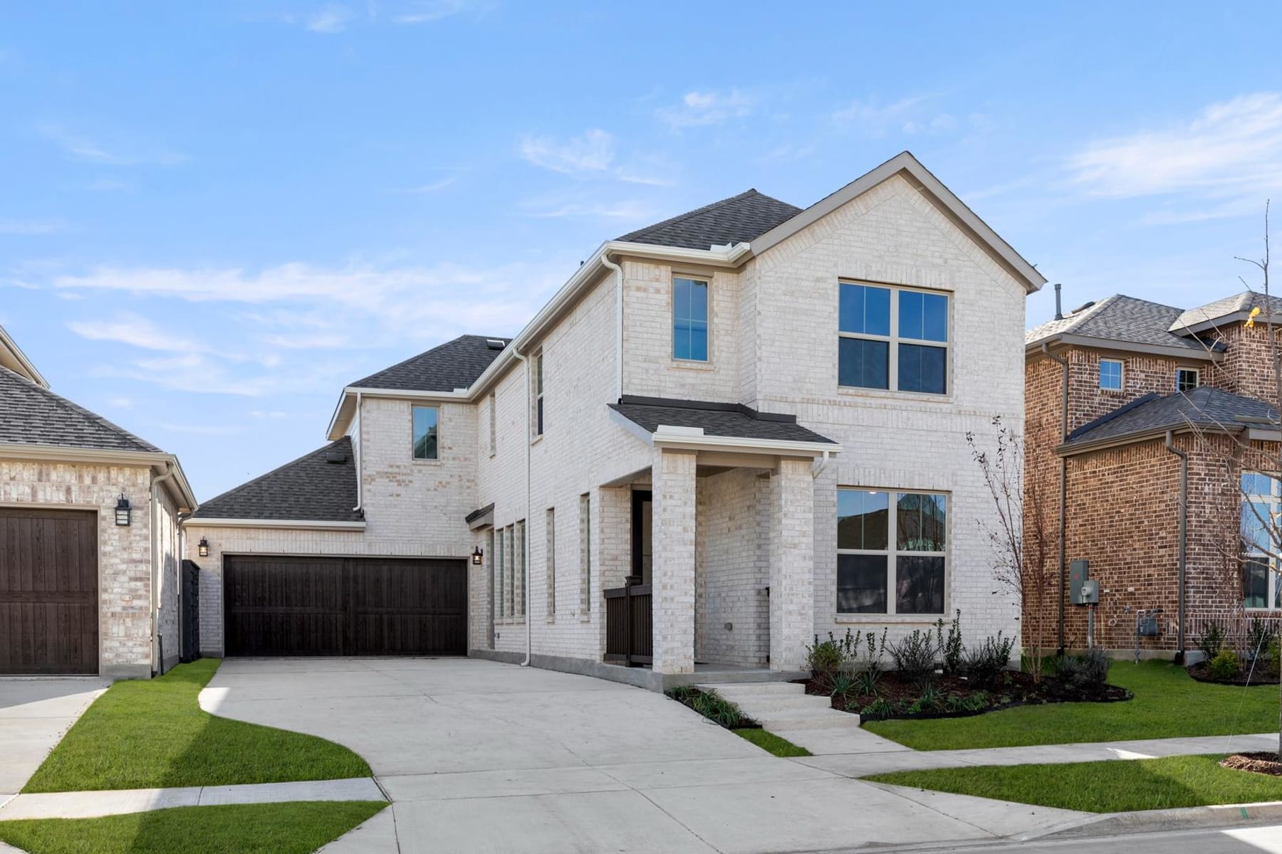 A two-story residential house with a garage, surrounded by a well-manicured lawn and a paved driveway, set against a clear blue sky with scattered clouds.
