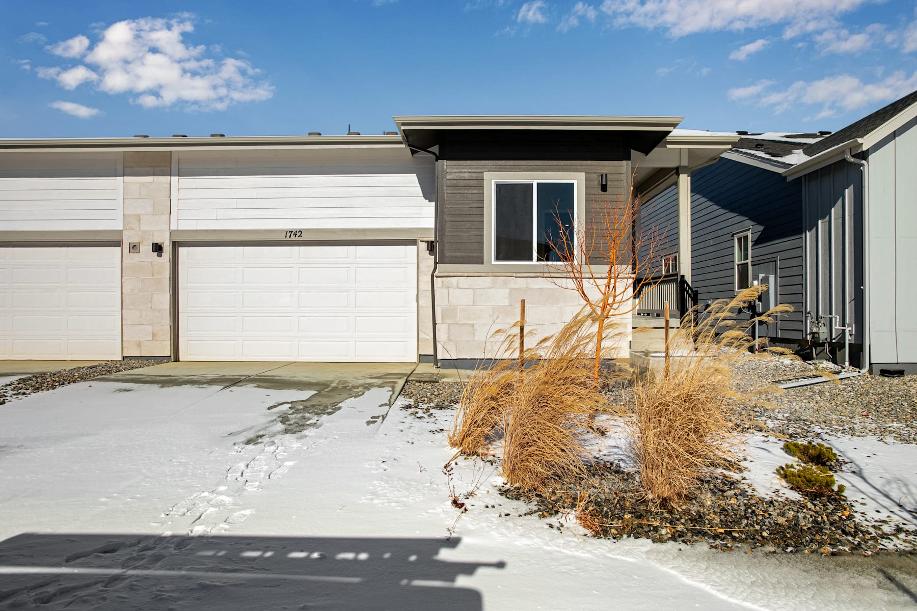 A modern, two-story residential building with a garage door and a snow-covered pathway leading to the entrance, set against a clear blue sky with scattered clouds.