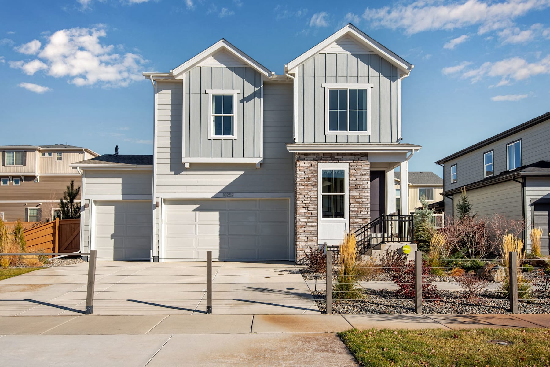 A modern two-story house with a gray exterior, stone accents, and a well-landscaped front yard set against a clear blue sky with scattered clouds.