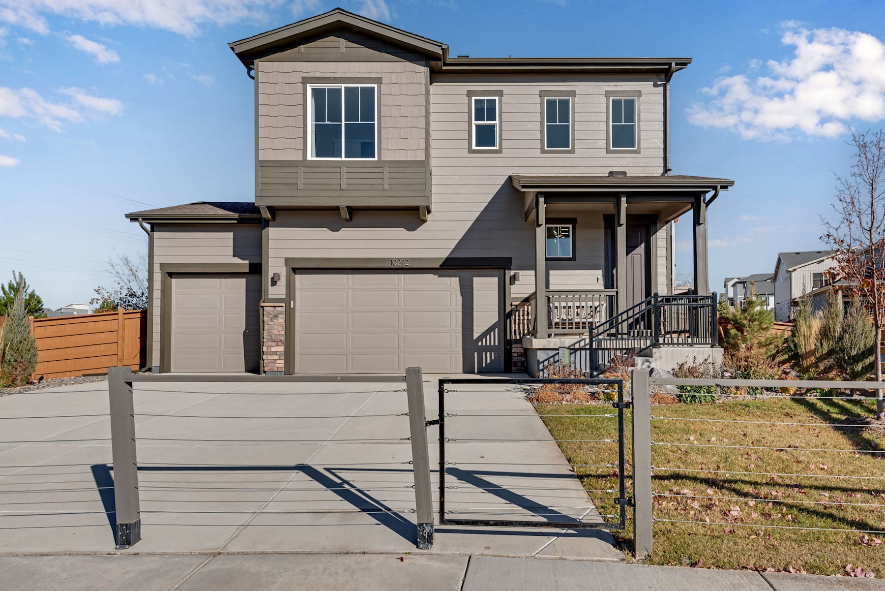 A two-story residential house with a gray exterior, a covered porch, and a fenced yard in the foreground, set against a backdrop of a clear sky with some clouds.