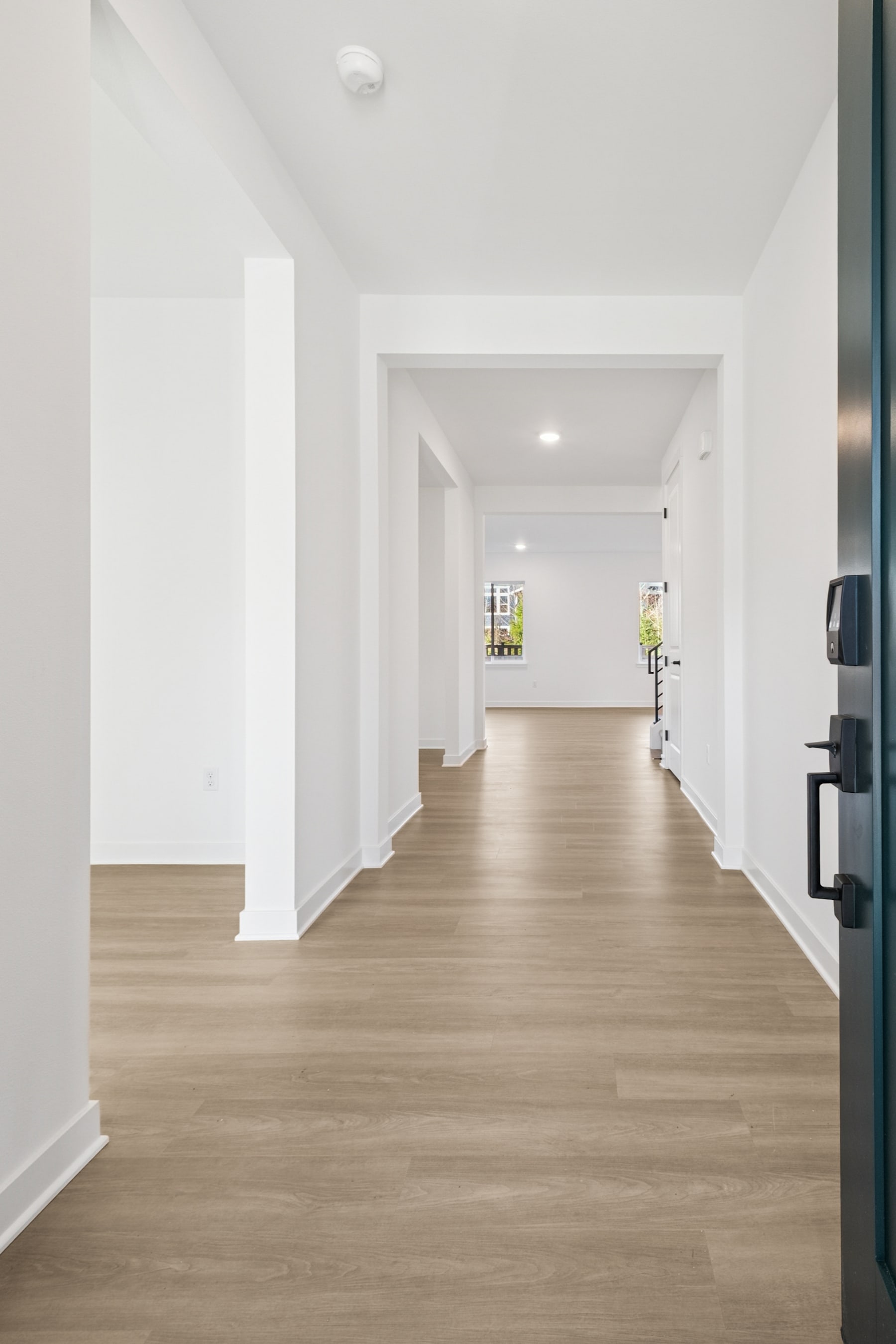 A spacious, minimalist hallway with light-colored hardwood floors and white walls, leading to a bright and airy space beyond.