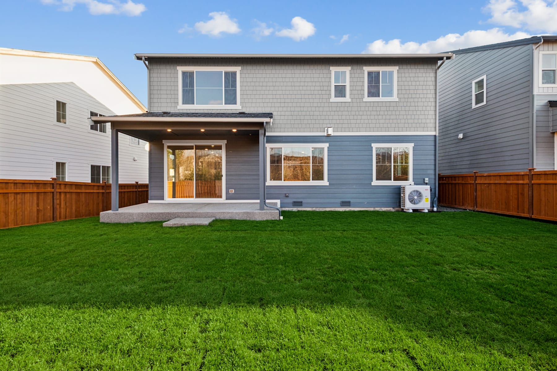 A two-story residential house with a covered porch, surrounded by a lush green lawn and a wooden fence in the foreground, set against a backdrop of a partly cloudy sky.