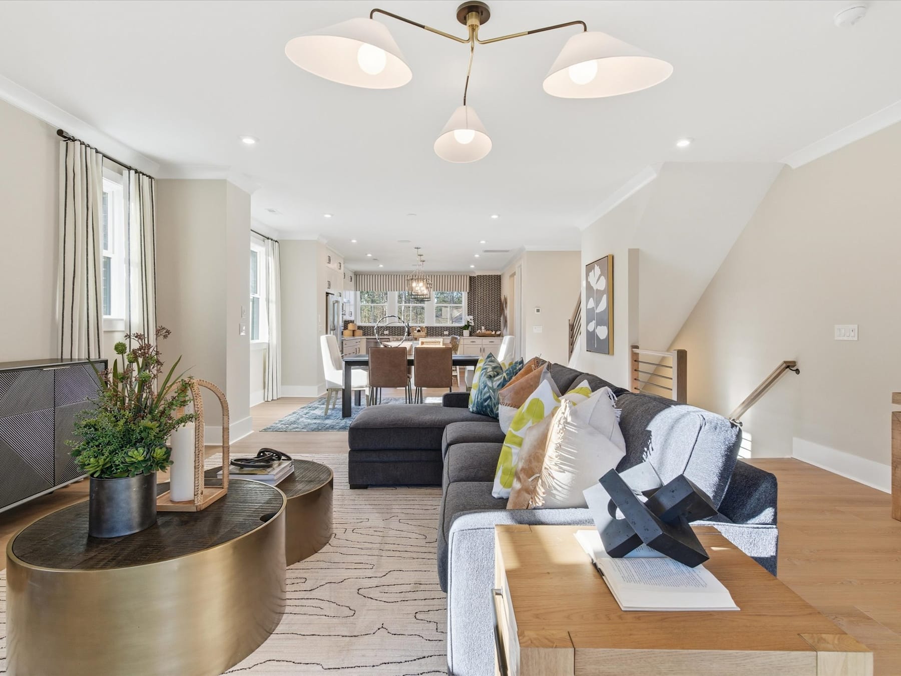 A modern and stylish living room with a gray sectional sofa, a wooden coffee table, and a chandelier with multiple light fixtures, leading into an open-concept kitchen and dining area.
