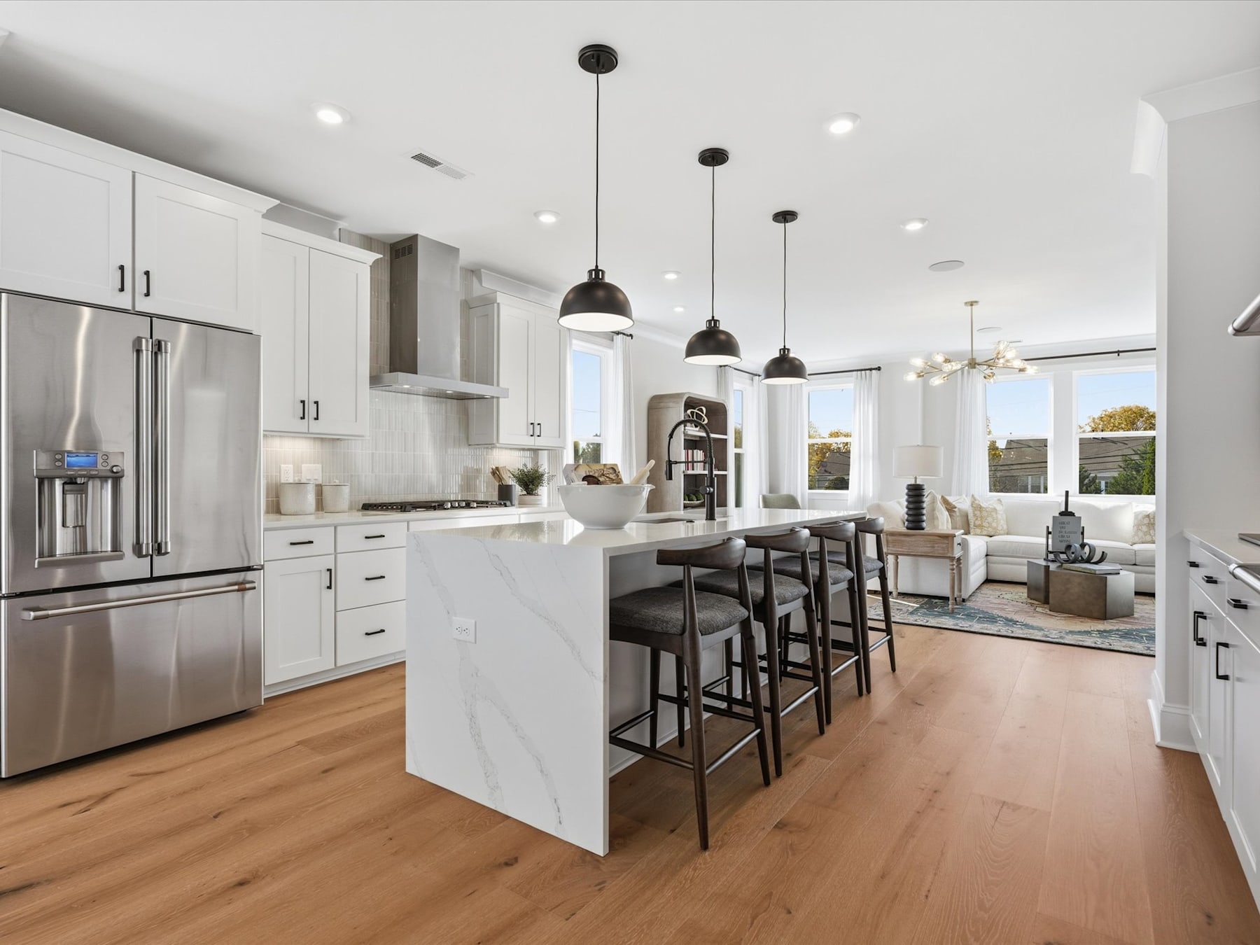 A modern, open-concept kitchen with white cabinets, stainless steel appliances, and a large island with bar stools, set against a backdrop of large windows overlooking a scenic outdoor view.
