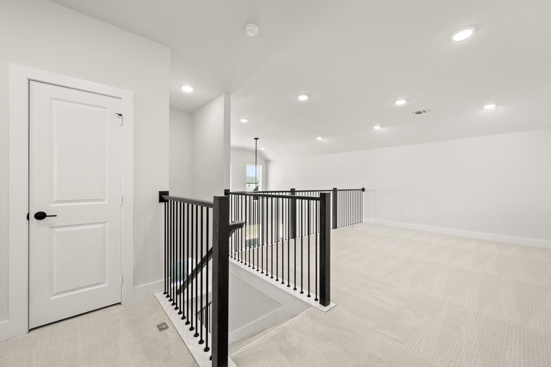 A bright, open hallway with a white door and a black metal railing leading up a set of stairs, surrounded by a clean, minimalist interior design.