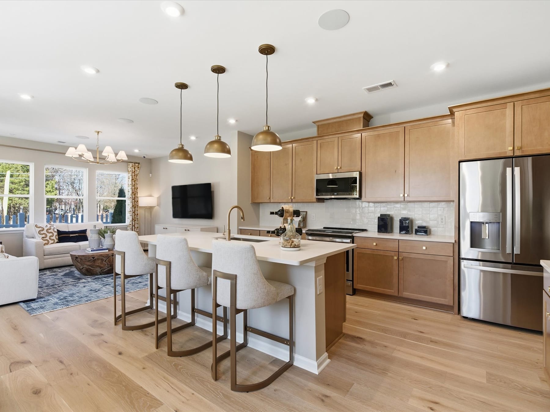 A modern and spacious kitchen with light wood cabinets, stainless steel appliances, and a central island with bar stools, surrounded by a cozy living room area with a large window overlooking the outdoors.