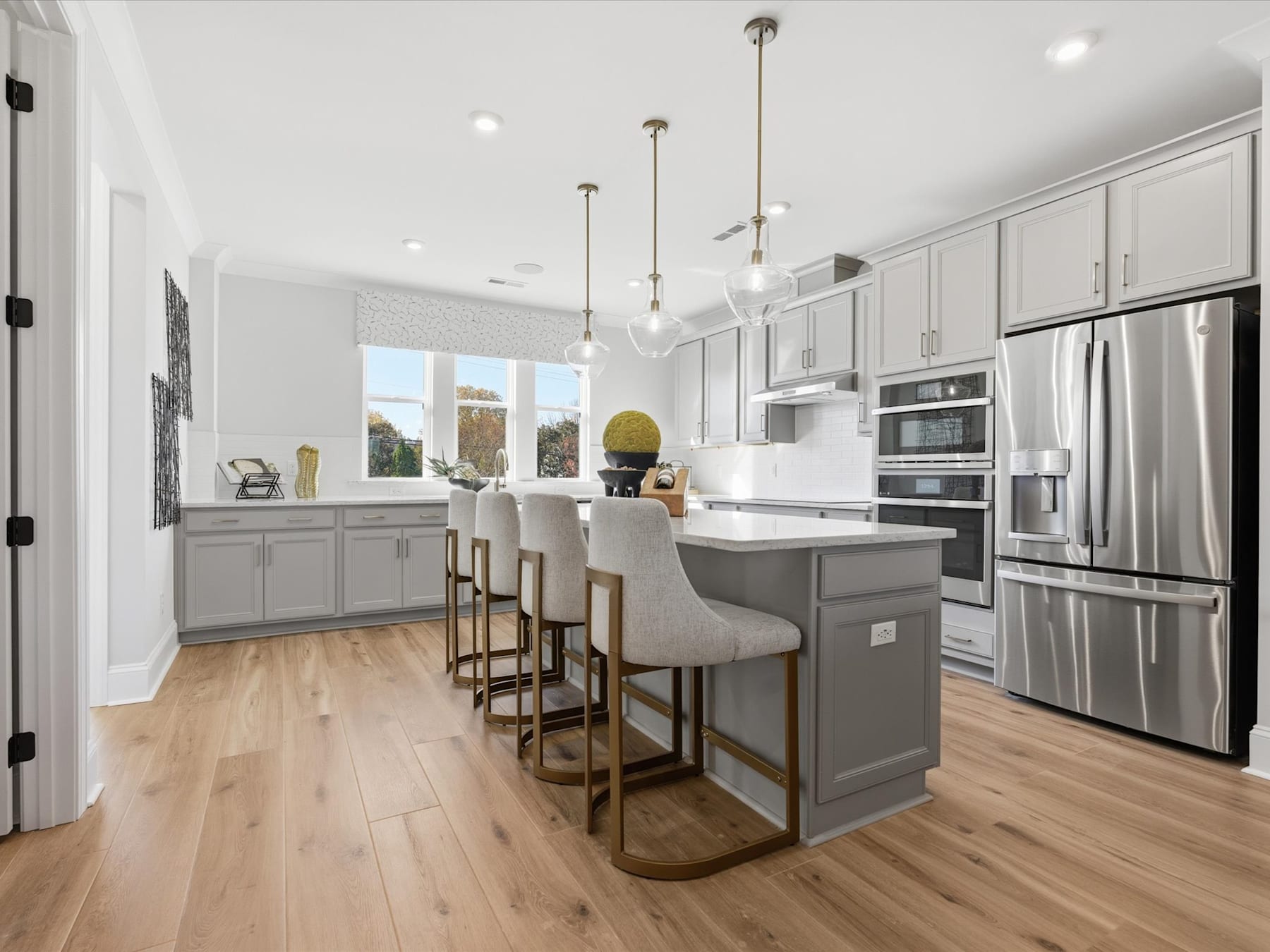 A modern, open-concept kitchen with white cabinets, gray island, stainless steel appliances, and hardwood floors, featuring pendant lighting and bar stools.