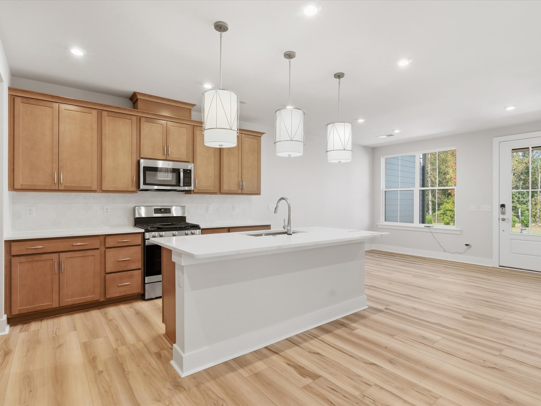 A modern, open-concept kitchen with light wood cabinets, a white island, and pendant lighting fixtures, set against a backdrop of hardwood floors and a sliding glass door leading to the outdoors.