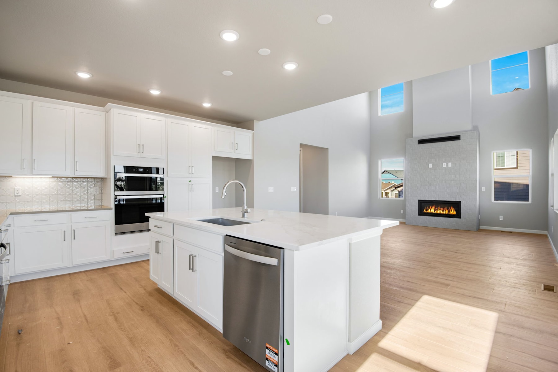 A modern, bright kitchen with white cabinets, stainless steel appliances, and a fireplace in the background, set against a backdrop of a clear sky visible through the windows.