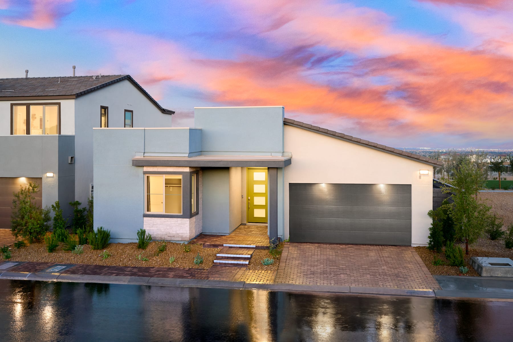 A modern, two-story house with a garage stands in the foreground, reflected in a body of water, against a vibrant, colorful sky at sunset.
