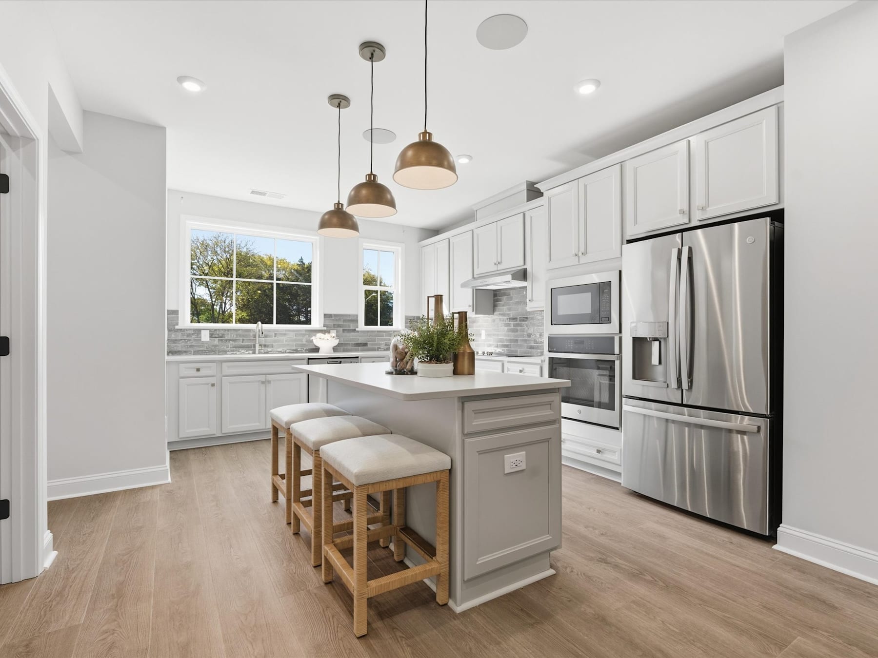 A modern, bright kitchen with white cabinets, stainless steel appliances, and a central island with stools, set against a backdrop of large windows overlooking a scenic outdoor view.