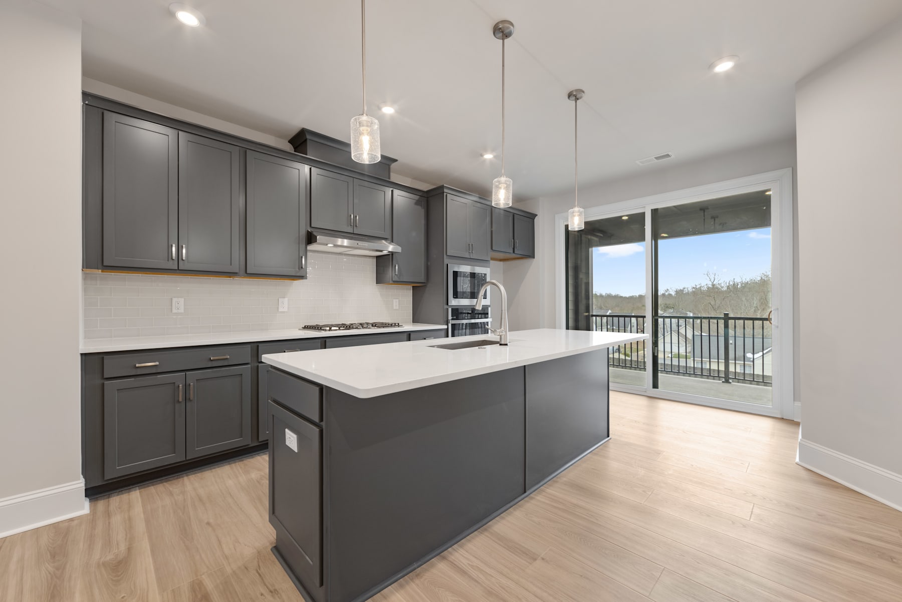 A modern, open-concept kitchen with dark gray cabinets, a white quartz countertop, and pendant lighting, overlooking a scenic outdoor view through a large window.