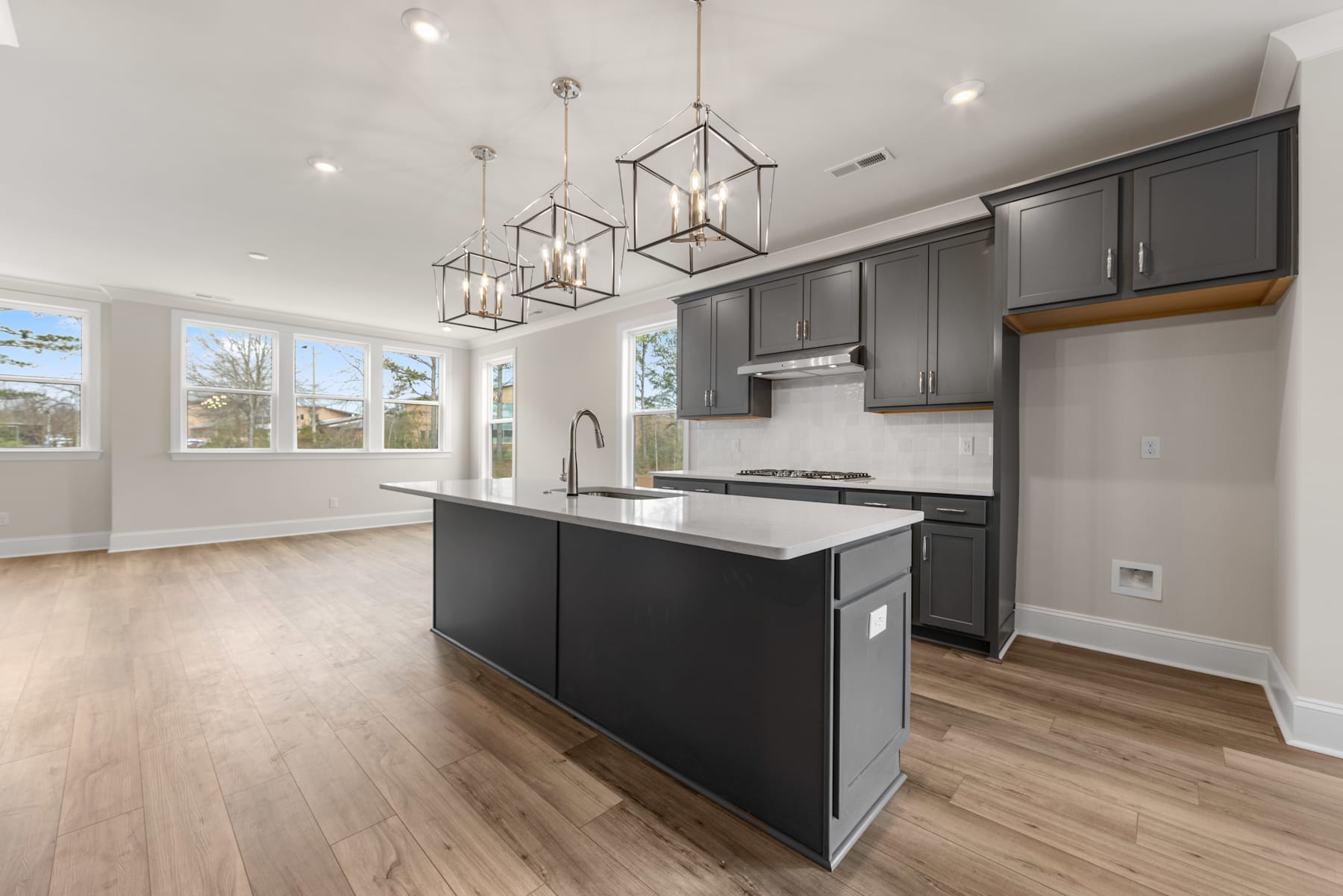 A modern and spacious kitchen with dark cabinets, a central island, and pendant lighting fixtures, set against a backdrop of large windows allowing natural light to flood the room.