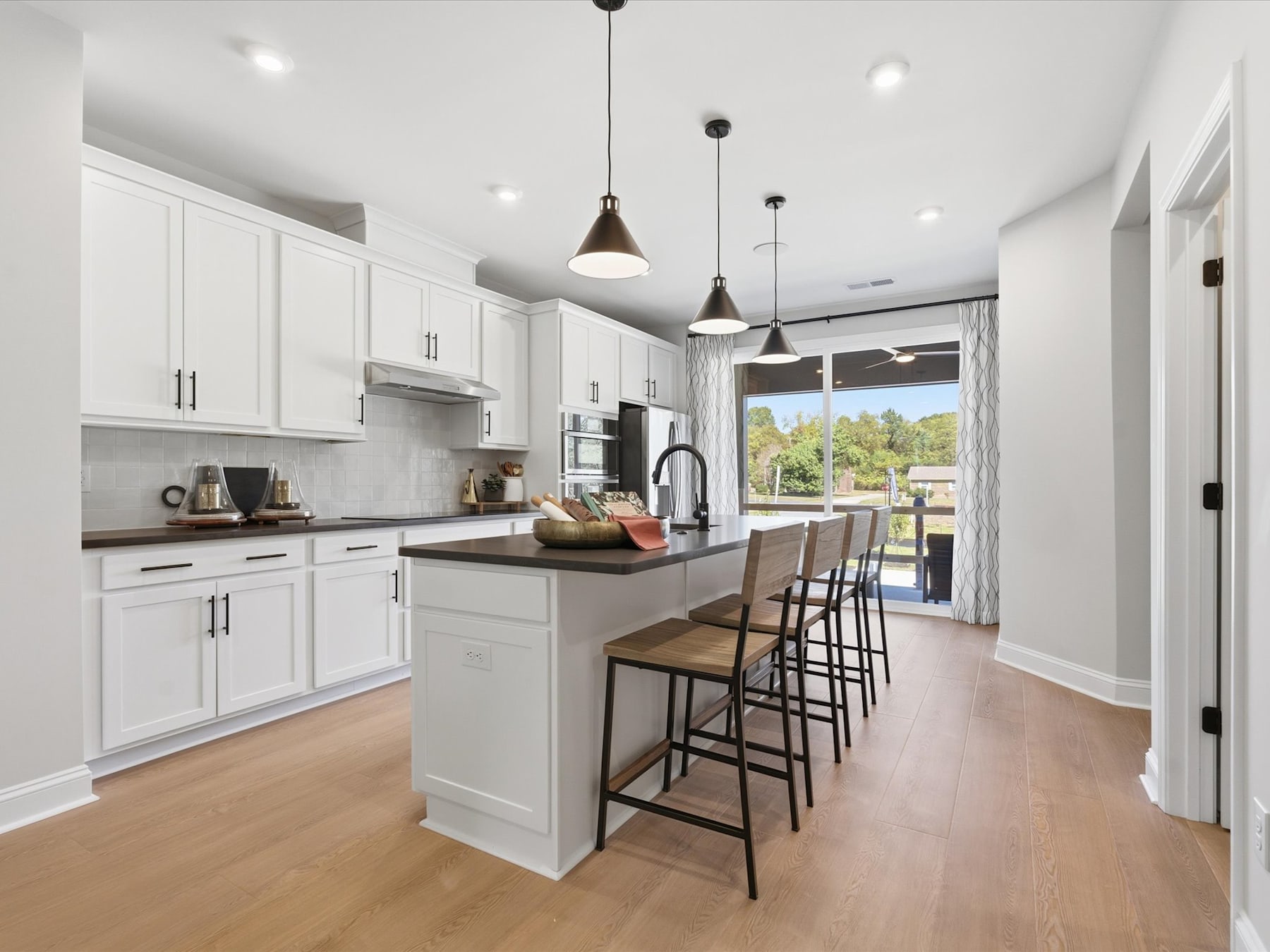 A modern, open-concept kitchen with white cabinets, a central island, and pendant lighting, set against a backdrop of a scenic outdoor view through a large window.