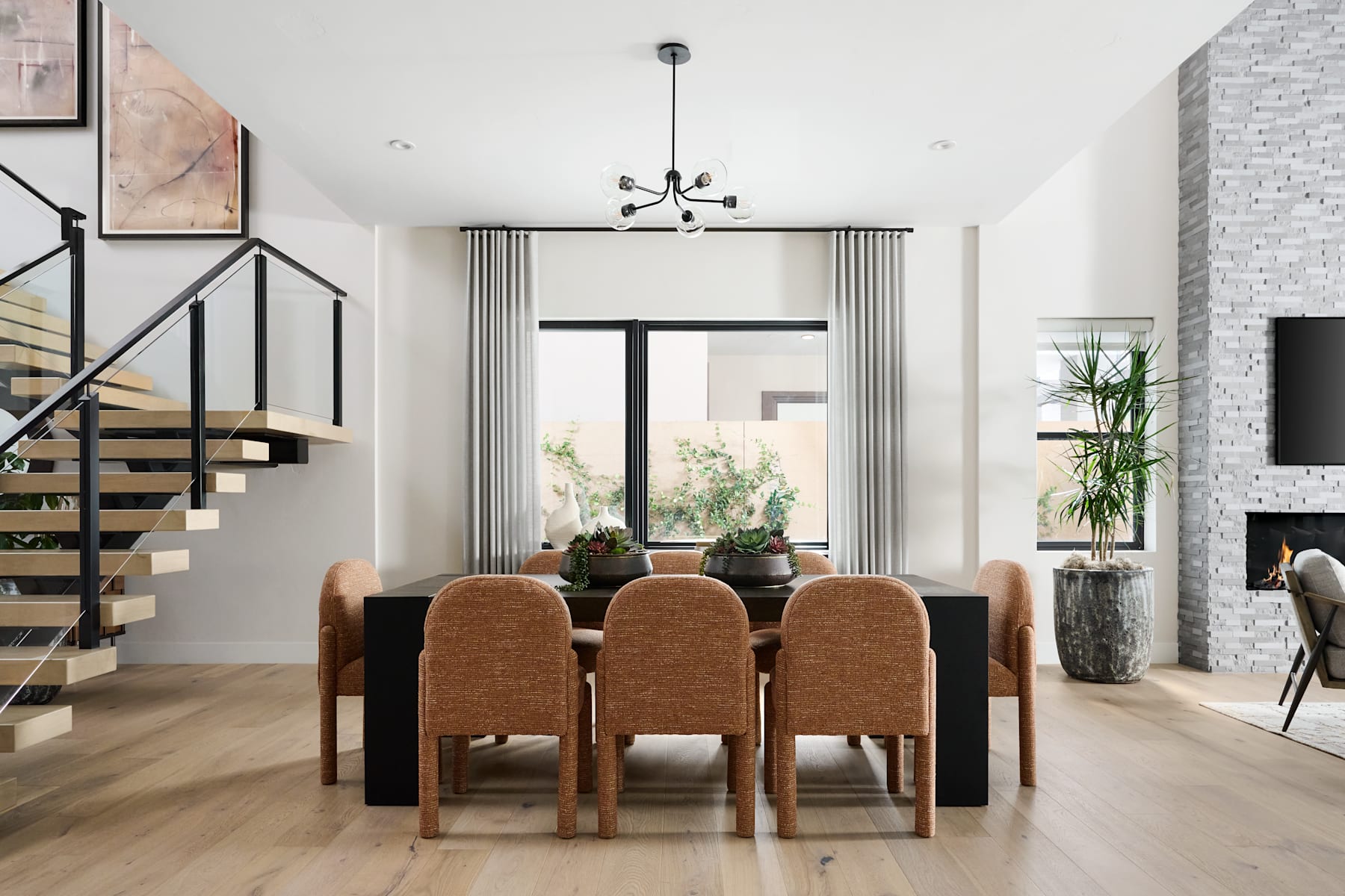 A modern and minimalist dining room with a wooden table and chairs, a large window overlooking a natural landscape, and a sleek staircase in the background.