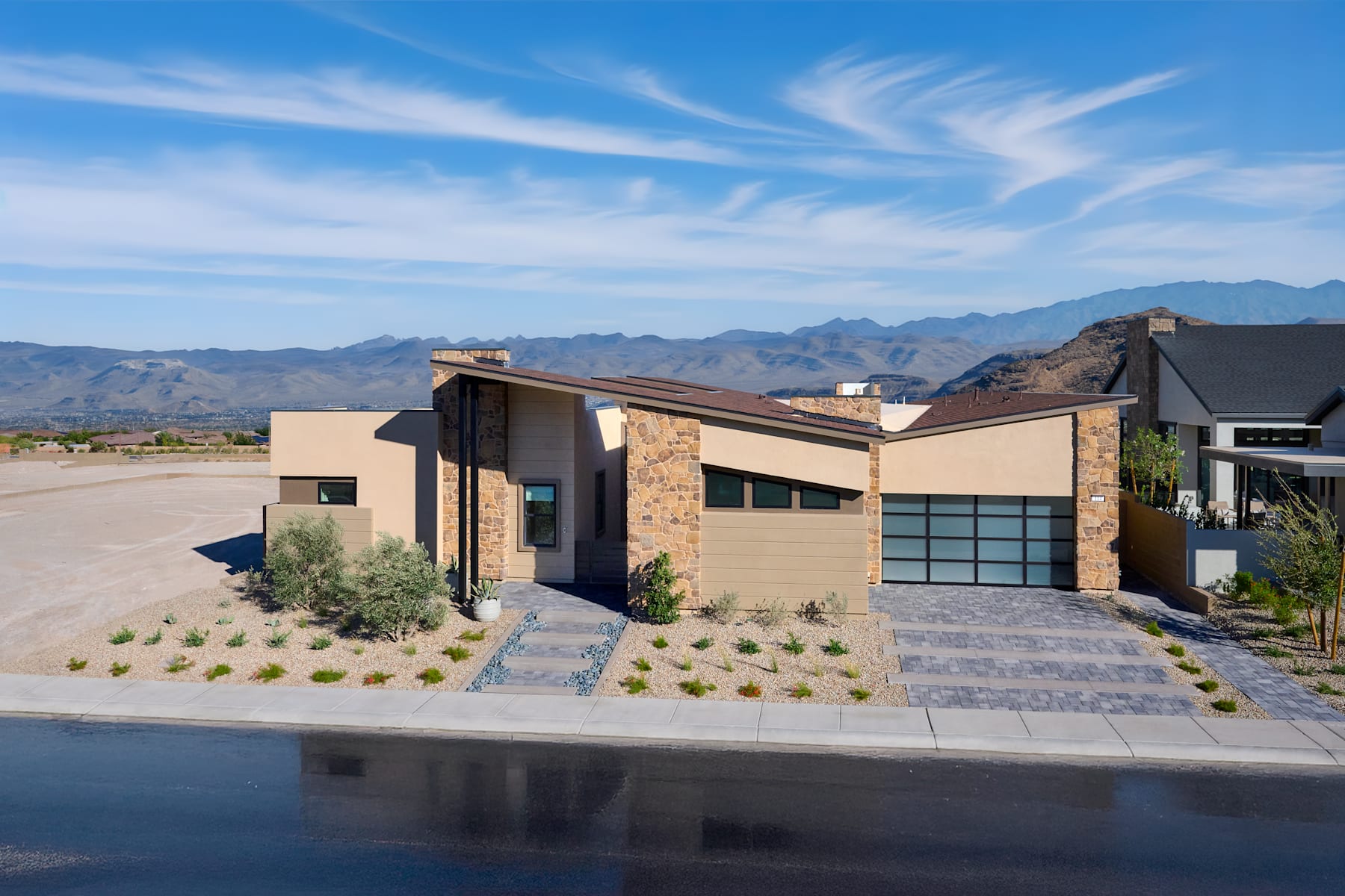 A modern, two-story house with a stone and stucco exterior, surrounded by a paved driveway and landscaped yard, set against a backdrop of mountains and a blue sky with wispy clouds.