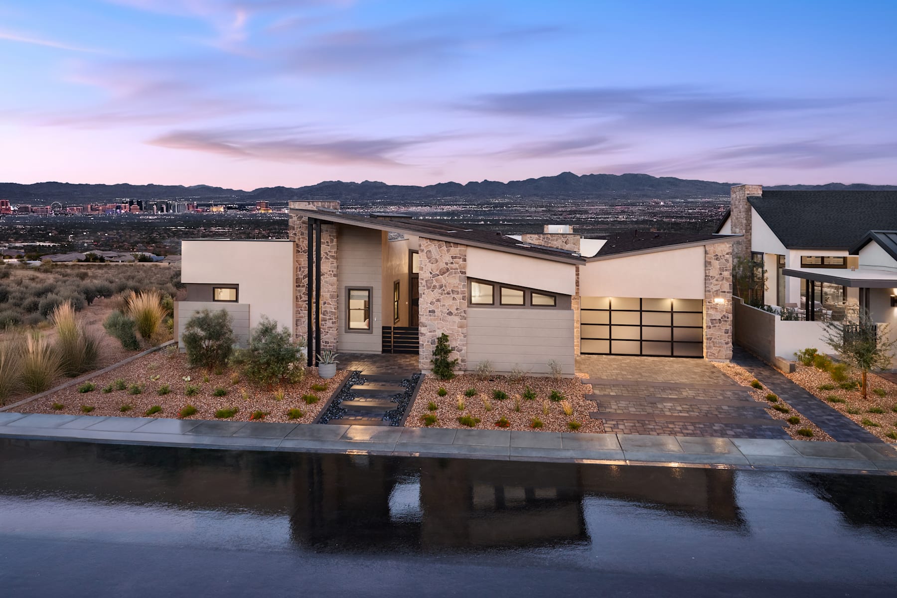 A modern, minimalist house with a stone and glass exterior sits atop a hill, overlooking a vast landscape of mountains and a vibrant sky at sunset.