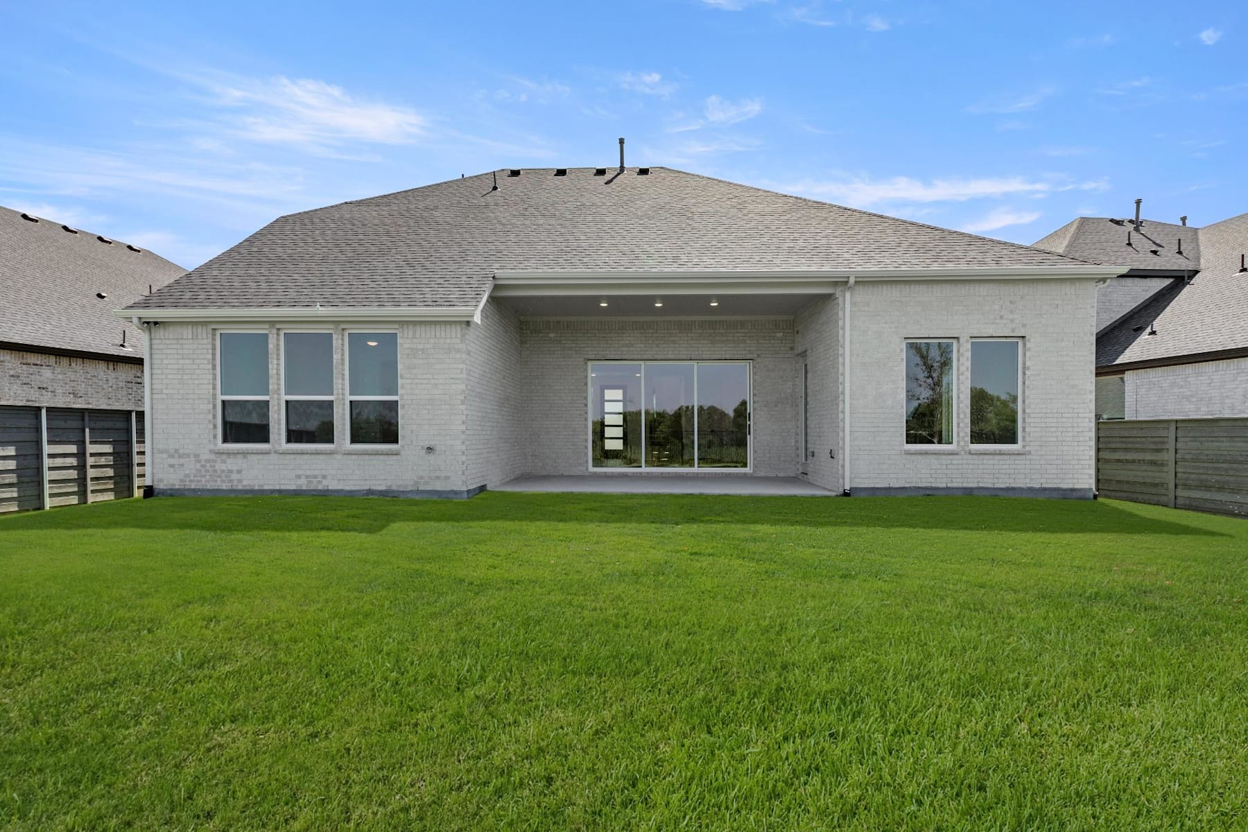 A modern, single-story house with a gray roof and large windows sits on a well-manicured lawn, surrounded by a clear blue sky with scattered clouds.
