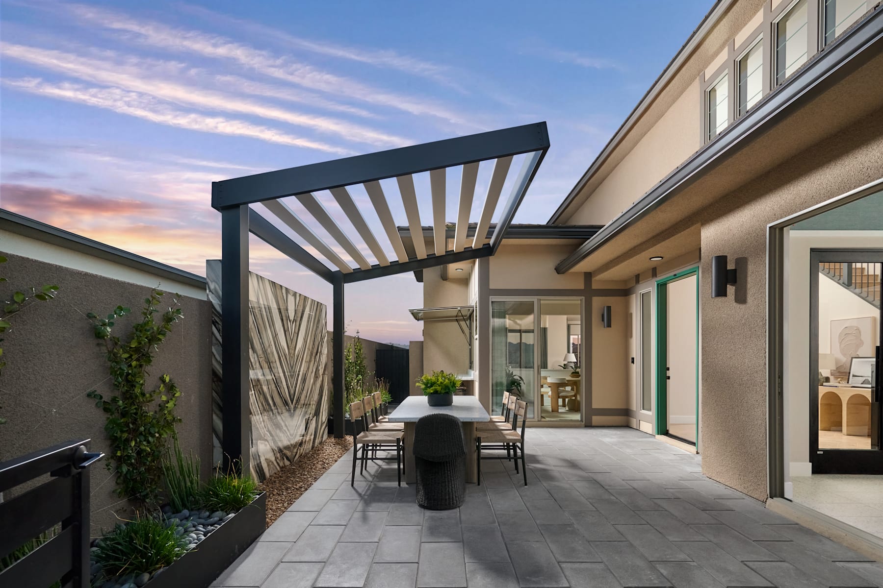 A modern outdoor patio area with a covered walkway, featuring a dining table and chairs, surrounded by lush greenery and a vibrant sunset sky.