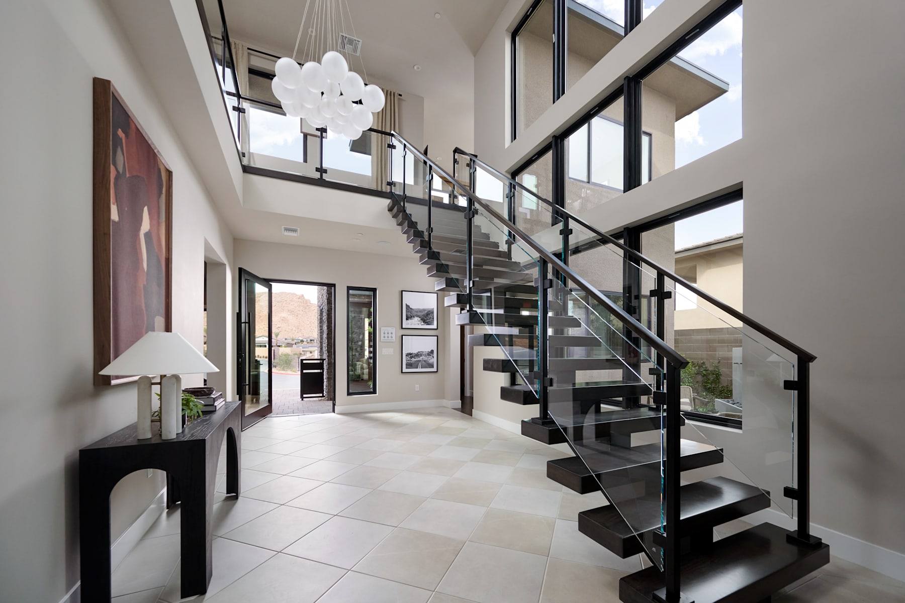A modern and spacious entryway with a sleek glass and metal staircase, a large chandelier, and a tiled floor leading to a room with a glass wall overlooking the exterior.