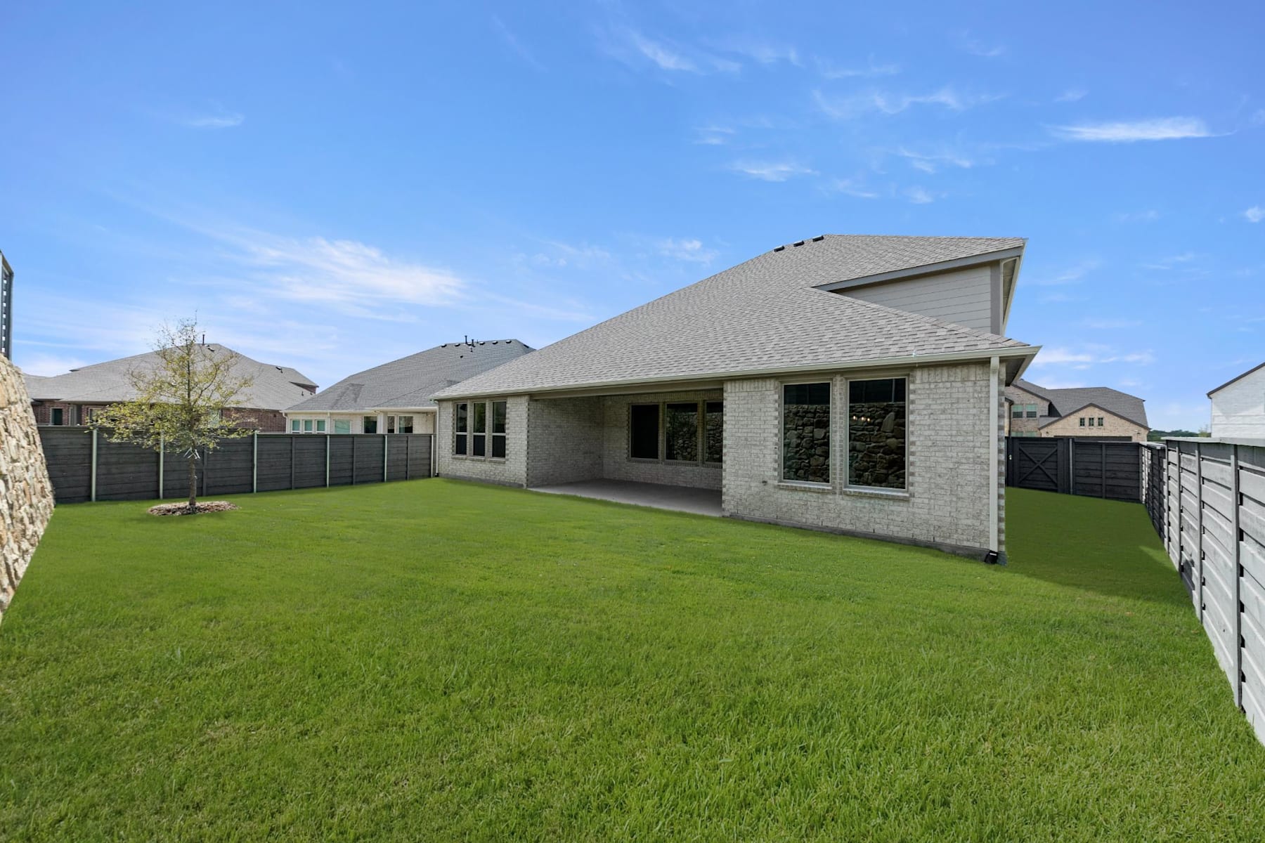 A well-manicured lawn surrounds a modern, two-story house with a gray roof and stone exterior, set against a clear blue sky with wispy clouds.